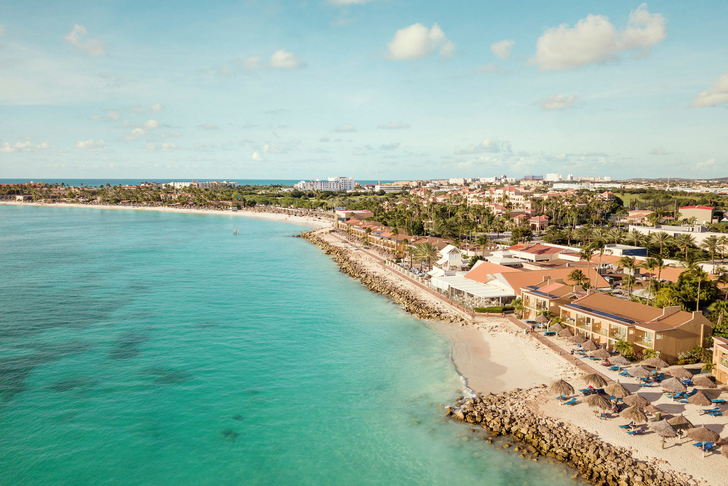 Strand med turkost vatten, omgivande byggnader och solparasoller. (AI-genererad text)