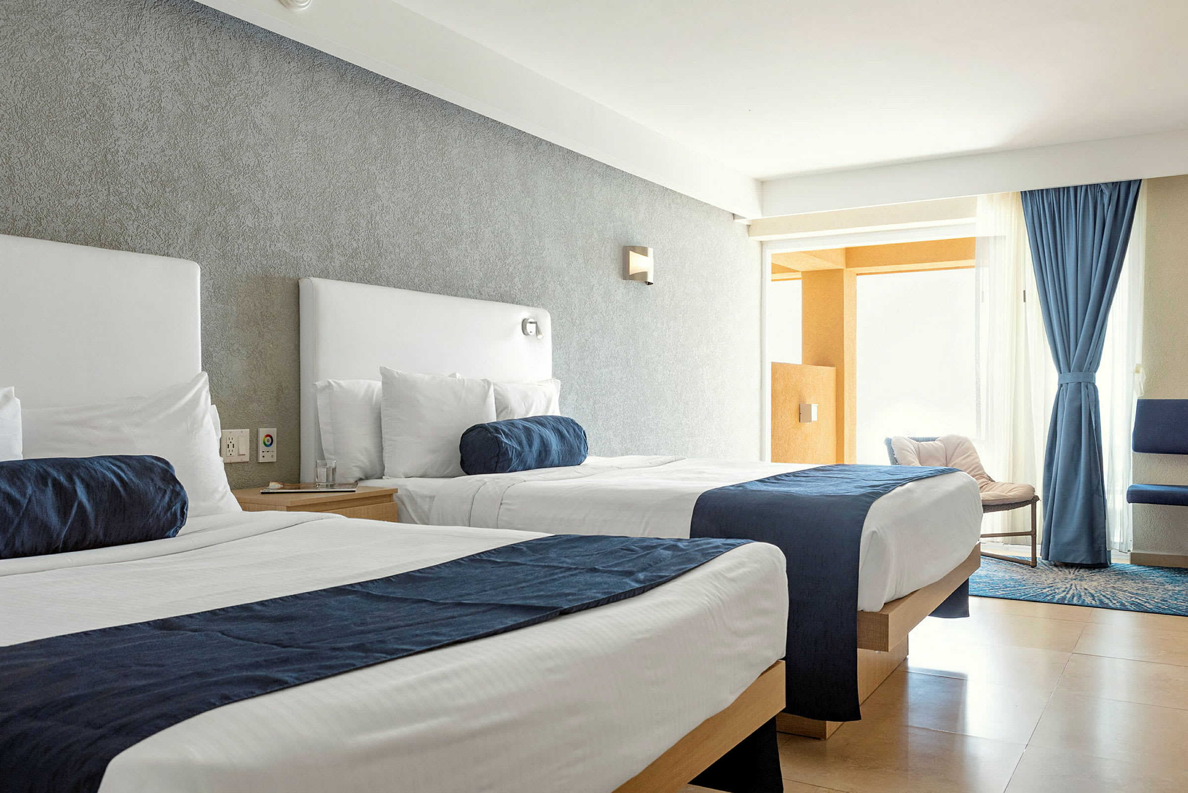 Ett hotellrum med två sängar med vita lakan och blå överkast. Fönstret är täckt med blå gardiner och ljus strömmar in i rummet. (AI-genererad text)