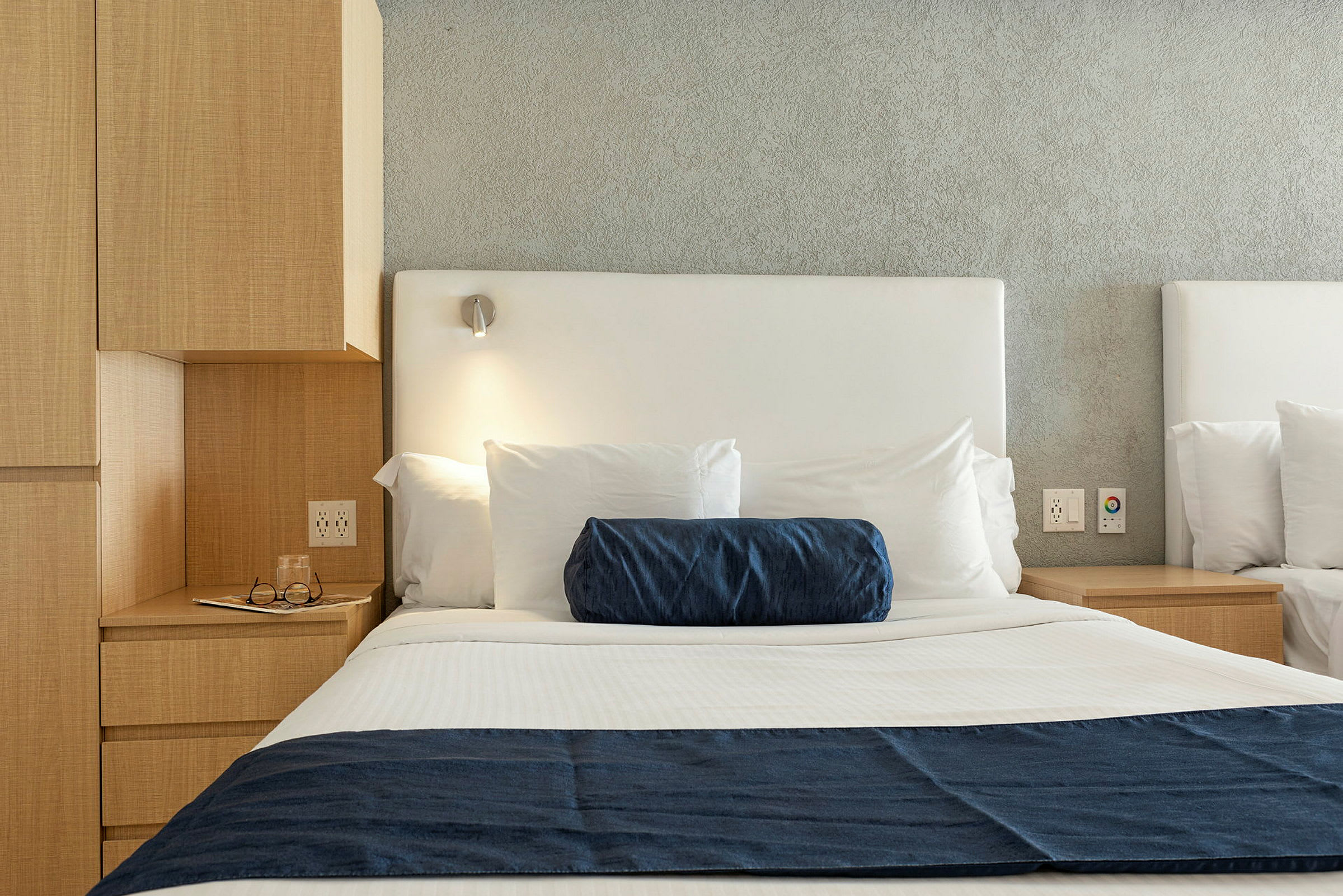 Bild på en hotellbäddning med vita lakan, en blå deko-kudde och en blå sängöverkast samt en sänglampa ovanför. (AI-genererad text)