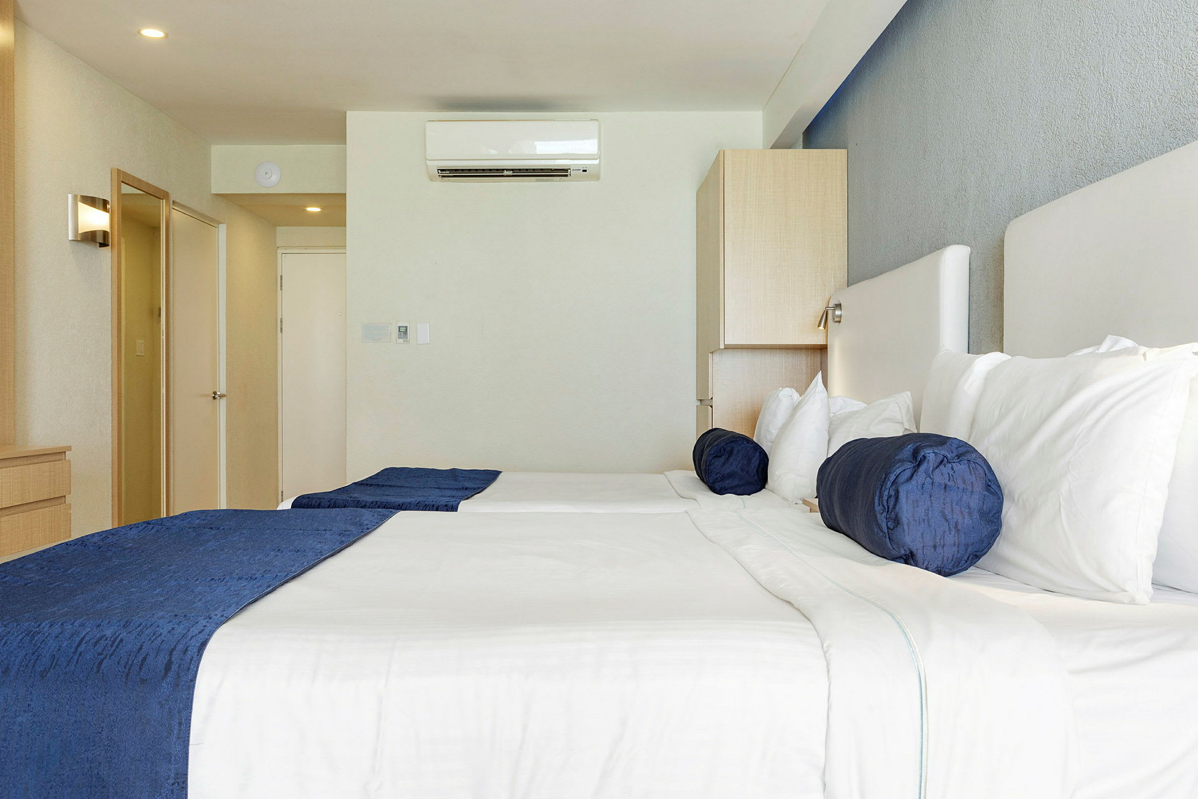 Ett hotellrum med två sängar, vita sängkläder och blå kuddar. En luftkonditioneringsenhet syns på väggen. (AI-genererad text)