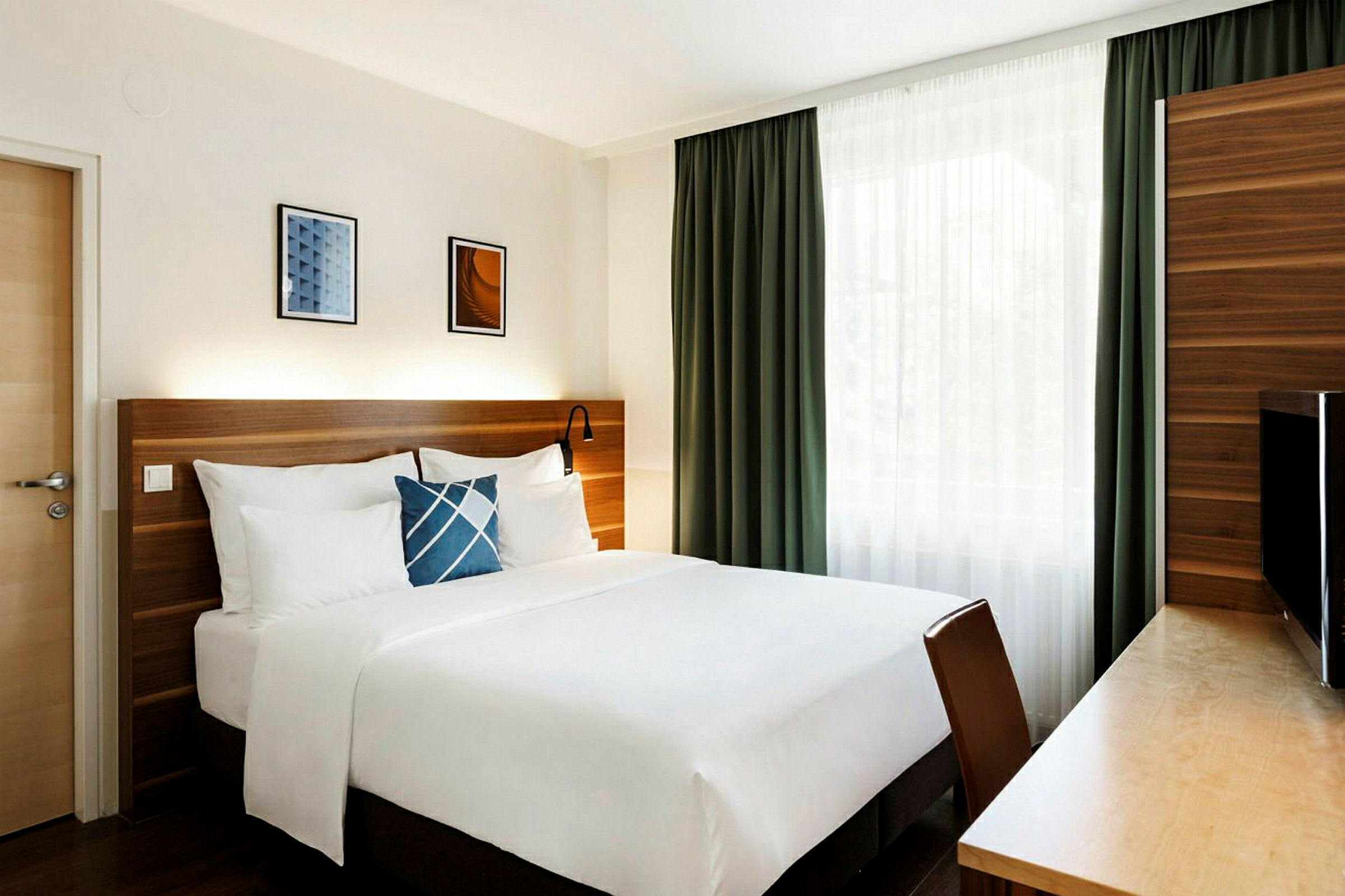 Ett hotellrum med en stor säng, vita sängkläder, en blå dekorationskudde, och en träpanel bakom. Gardiner hänger framför ett fönster. (AI-genererad text)