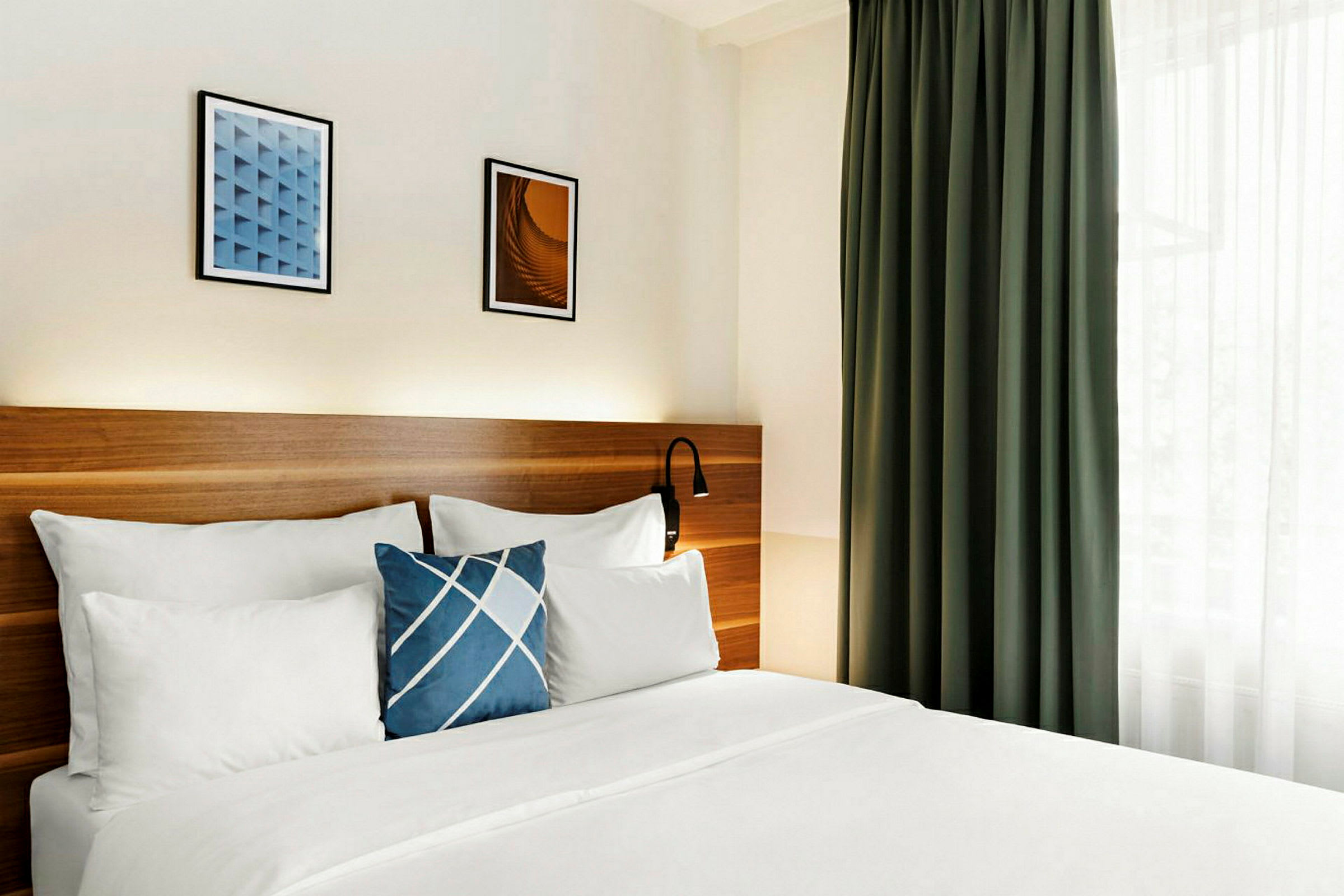 Ett hotellrum med en dubbelsäng, vita sängkläder och en blå kudde med mönster. Bakom sängen finns en träpanel och på väggen hänger två tavlor. Till höger syns ett grönt draperi. (AI-genererad text)