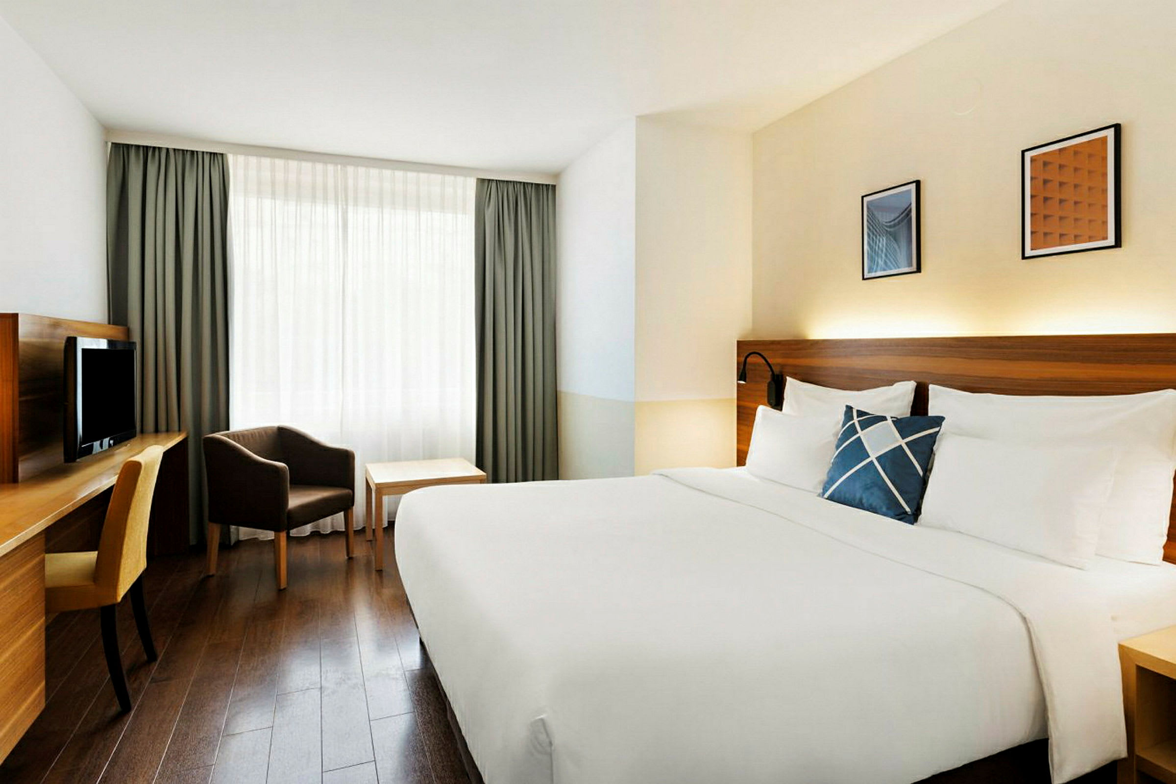 Ett hotellrum med en stor säng med vita sängkläder, ett skrivbord med stol, en fåtölj och ett bord vid ett fönster med gardiner. (AI-genererad text)