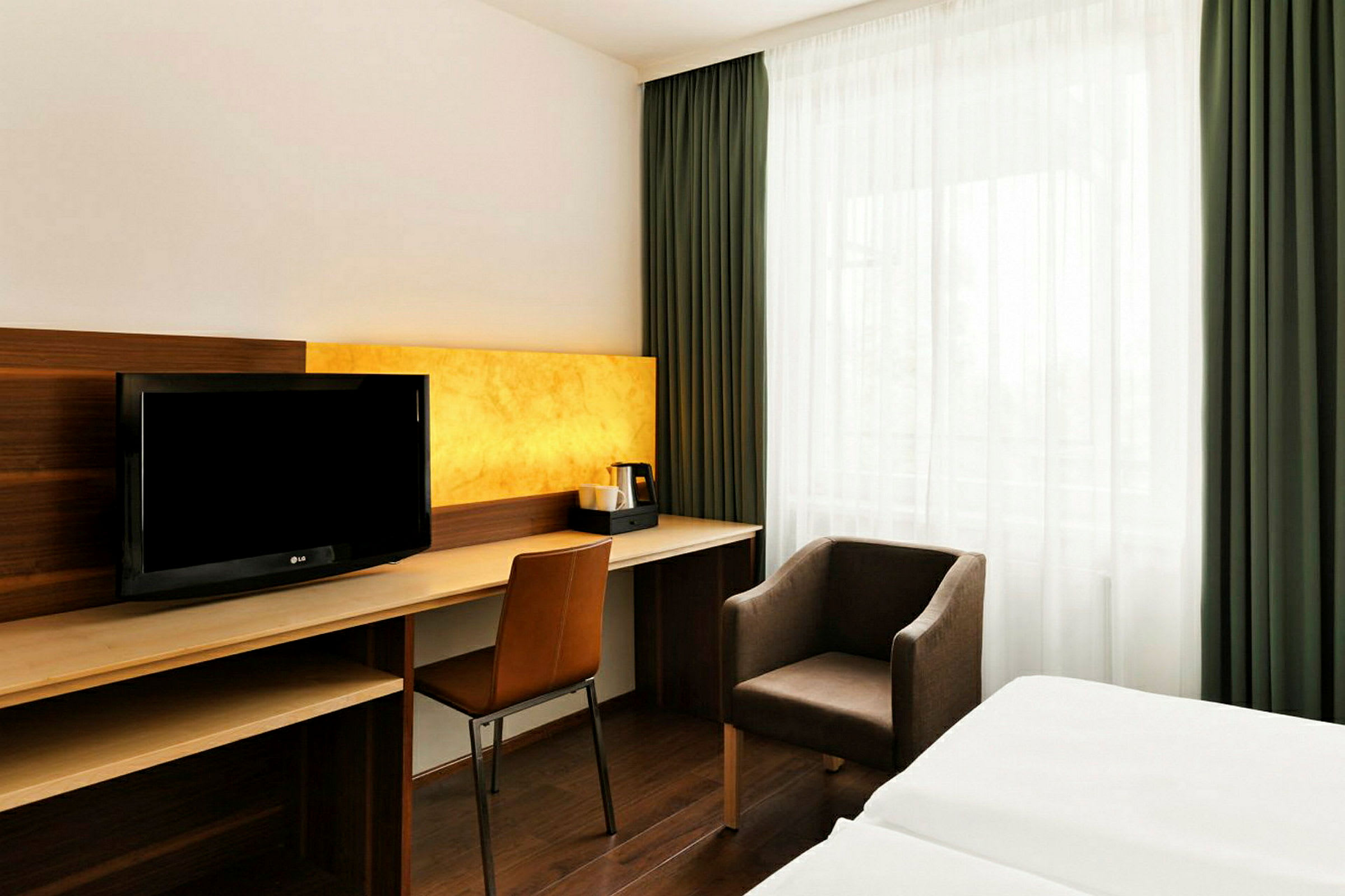 Ett hotellrum med skrivbord, stol, fåtölj och TV. Sängen syns delvis i bild och fönstret är täckt med gröna gardiner. (AI-genererad text)