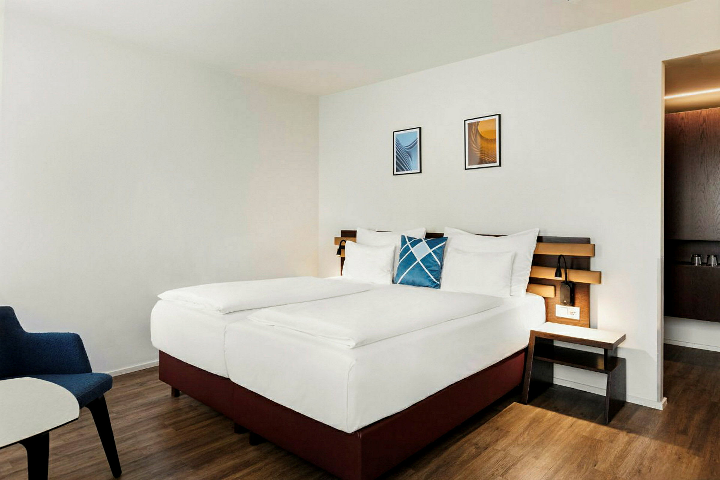 Hotellrum med en dubbelsäng, vita sängkläder och ett blått dekorativt kudde. En träpanel tjänar som sänggavel och rummet har trägolv. (AI-genererad text)