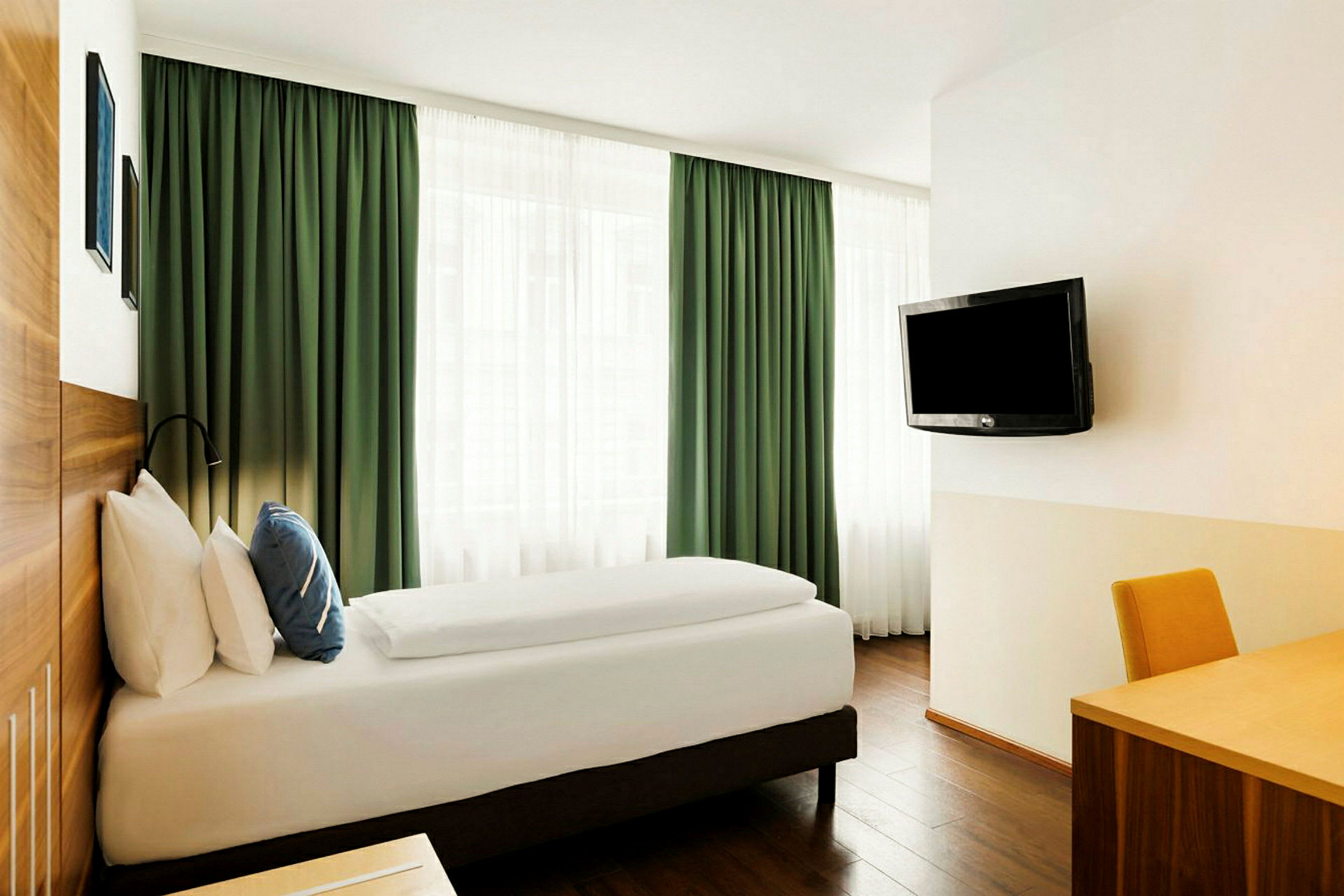 Ett hotellrum med en enkelsäng, gröna gardiner, ett skrivbord och en väggmonterad TV. (AI-genererad text)