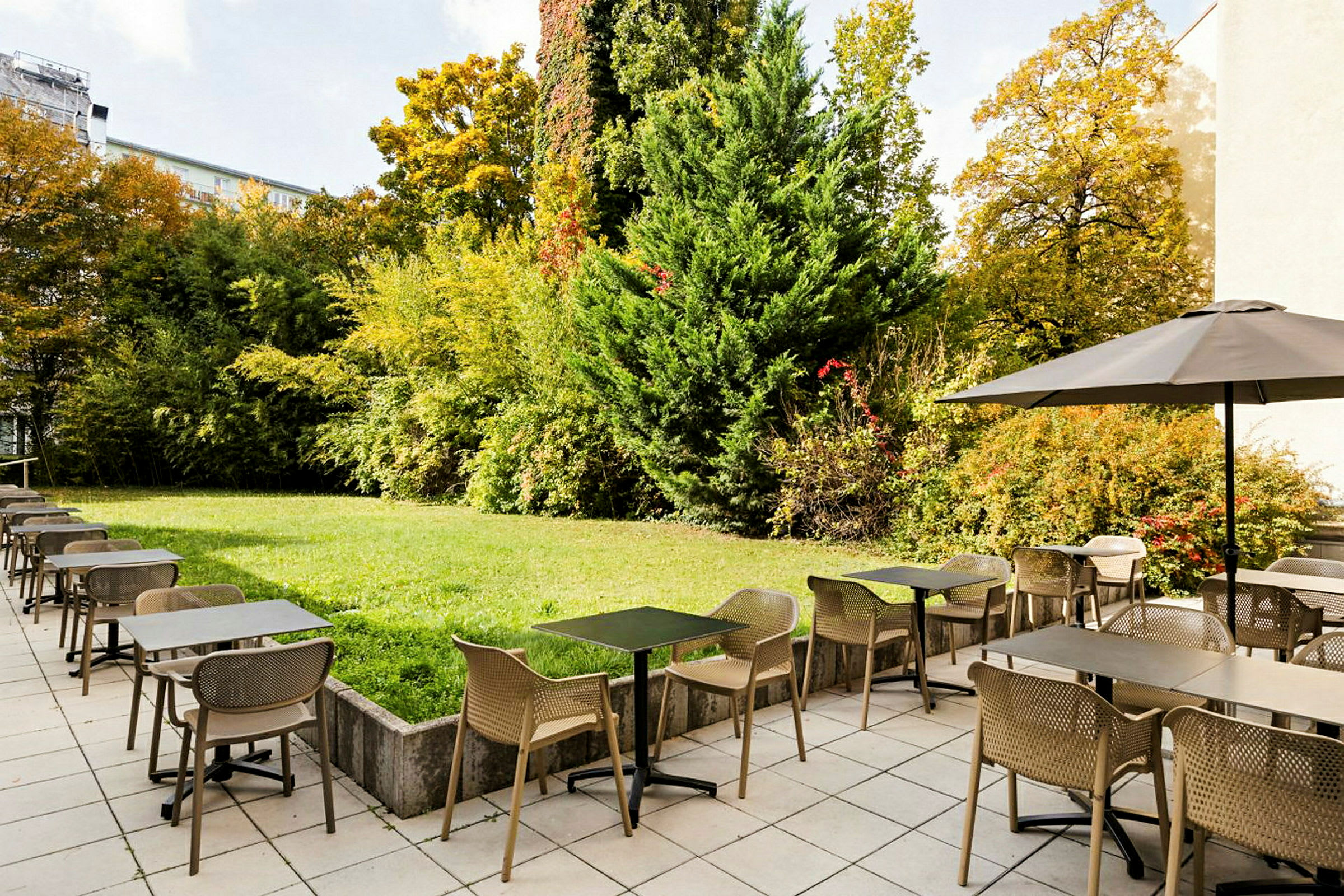 En utomhusrestaurang med bord och stolar på ett stenlagt område bredvid en grön trädgård med buskar och träd. (AI-genererad text)