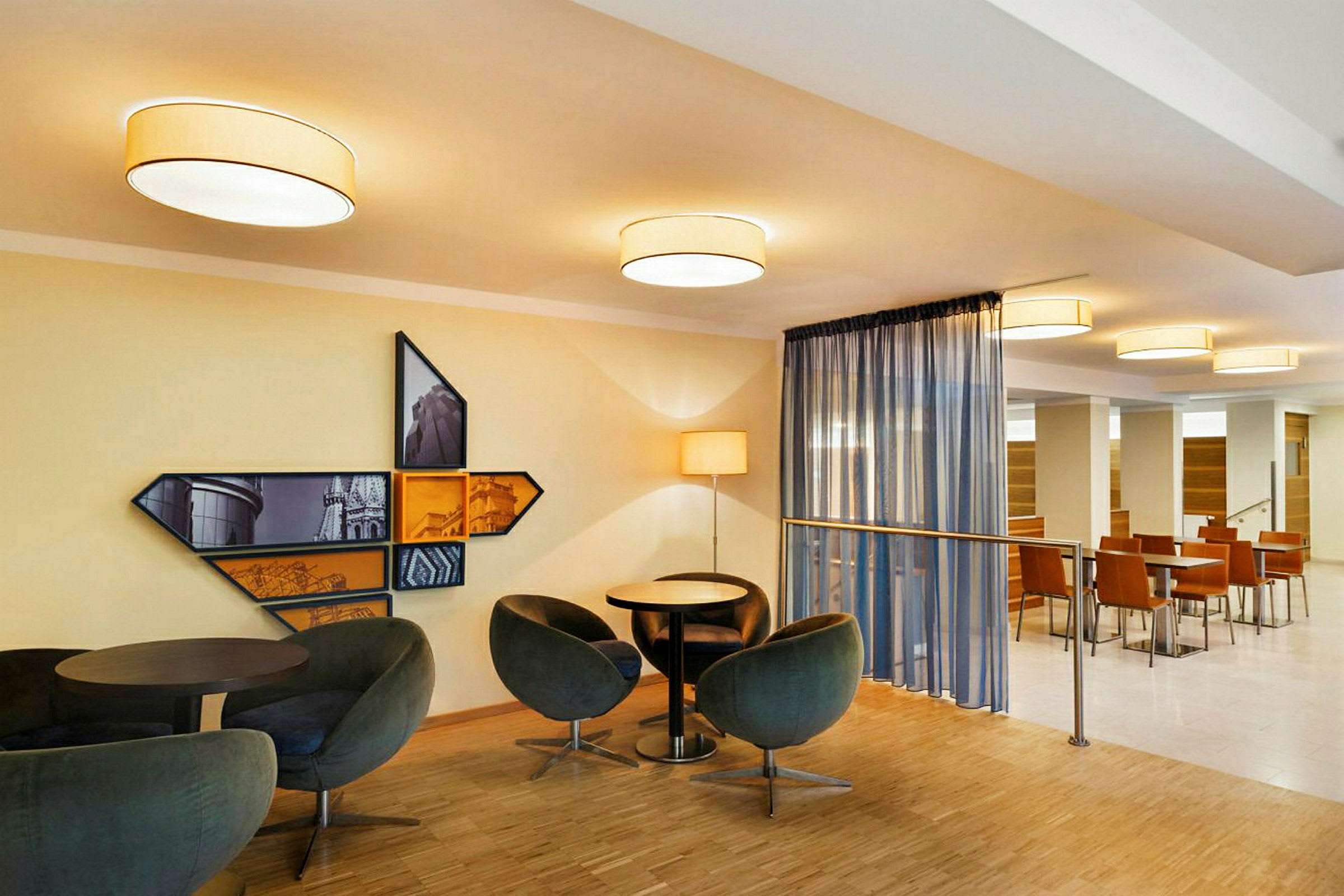 Ett modernt sällskapsrum med sittgrupper, runda bord och designade fåtöljer. På väggen finns en dekorativ tavla med geometriska ramar och stadsmotiv. Lamparmaturer ger en varm belysning. (AI-genererad text)