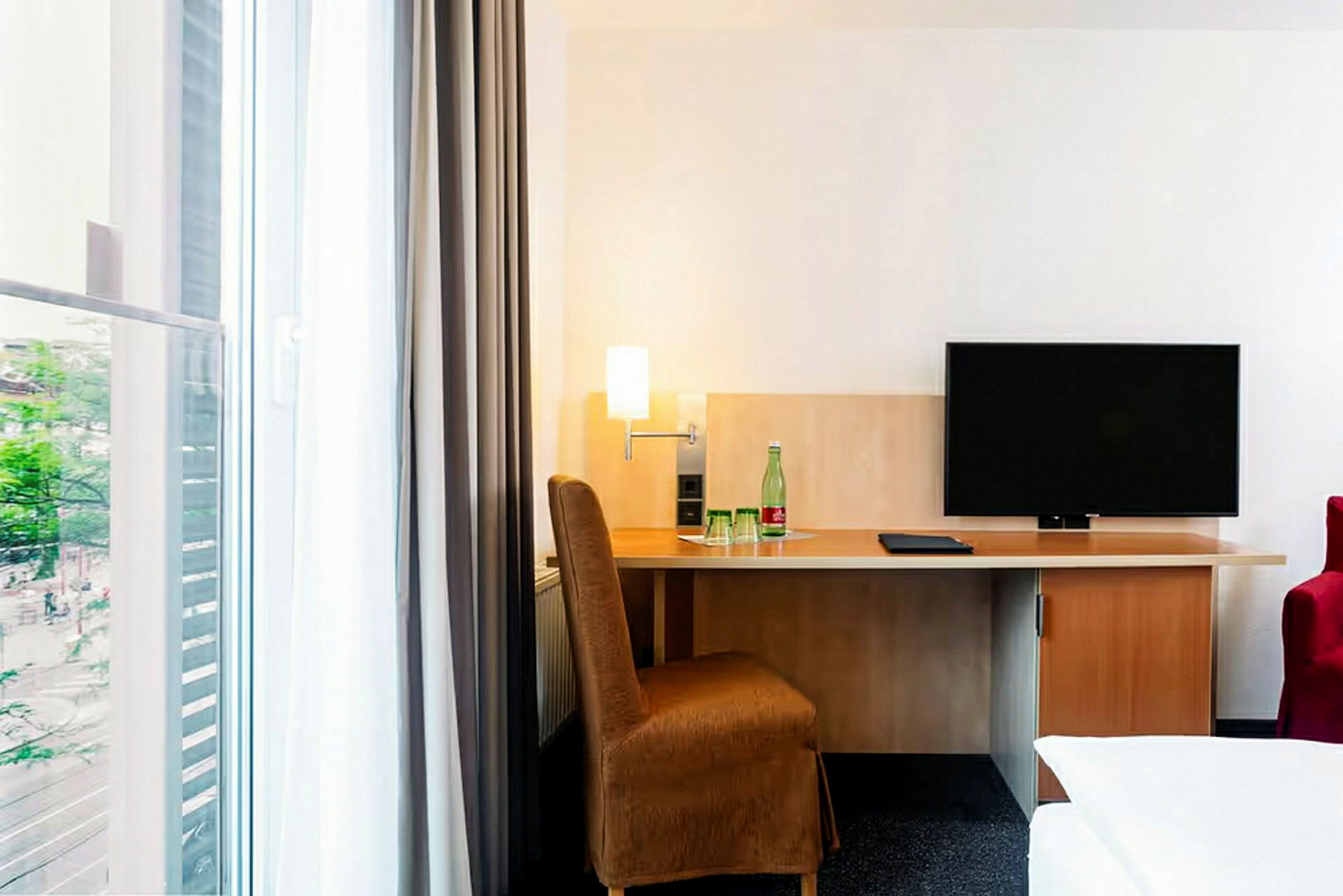 Ett hotellrum med ett skrivbord, stol och en TV. Ett fönster till vänster med gardiner släpper in ljus. (AI-genererad text)