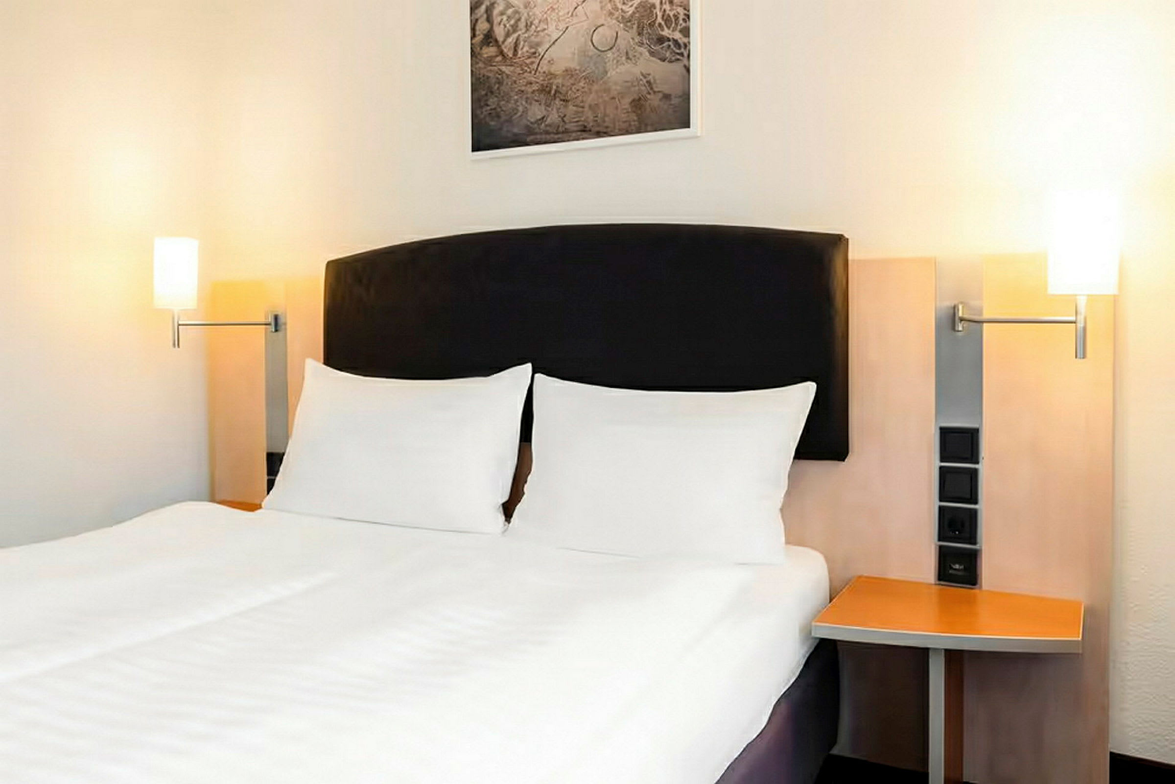 Ett hotellrum med en dubbelsäng, vita sängkläder och en mörk sänggavel. På båda sidor av sängen finns sängbord med lampor monterade på väggen. (AI-genererad text)