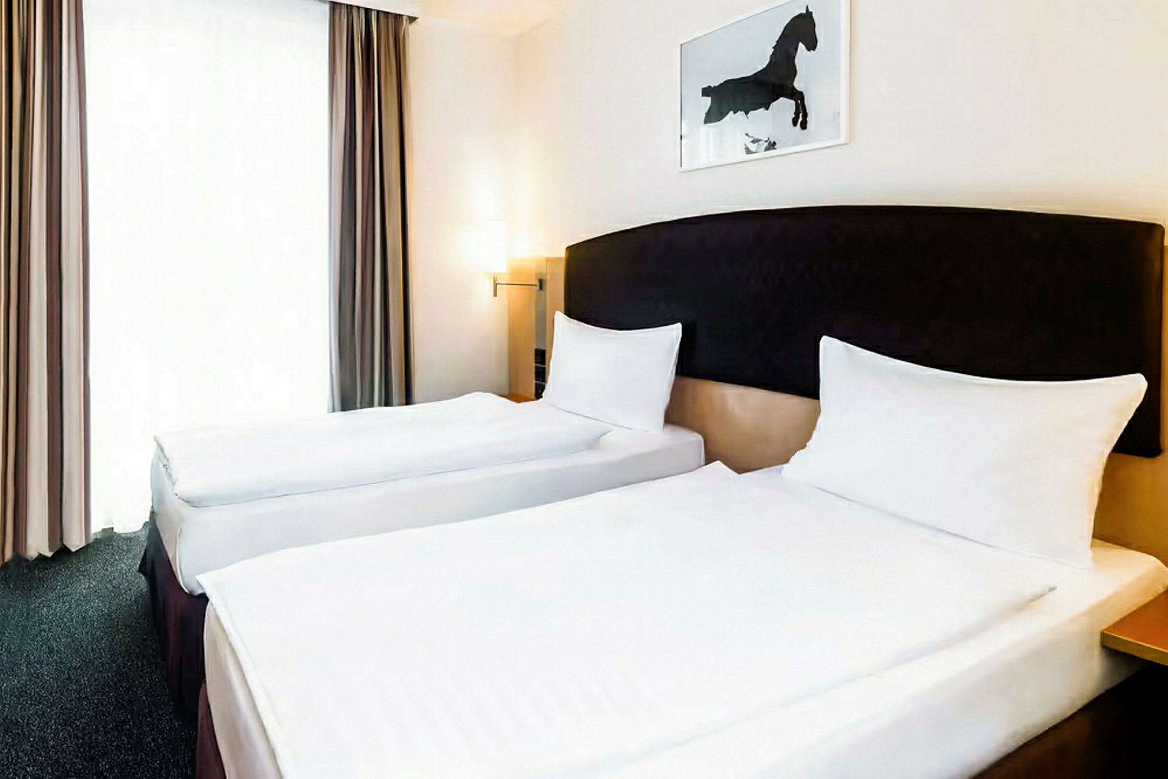 Två enkelsängar står bredvid varandra i ett hotellrum. Väggarna är ljusa och det finns ett konstverk av en häst ovanför huvudgaveln. (AI-genererad text)
