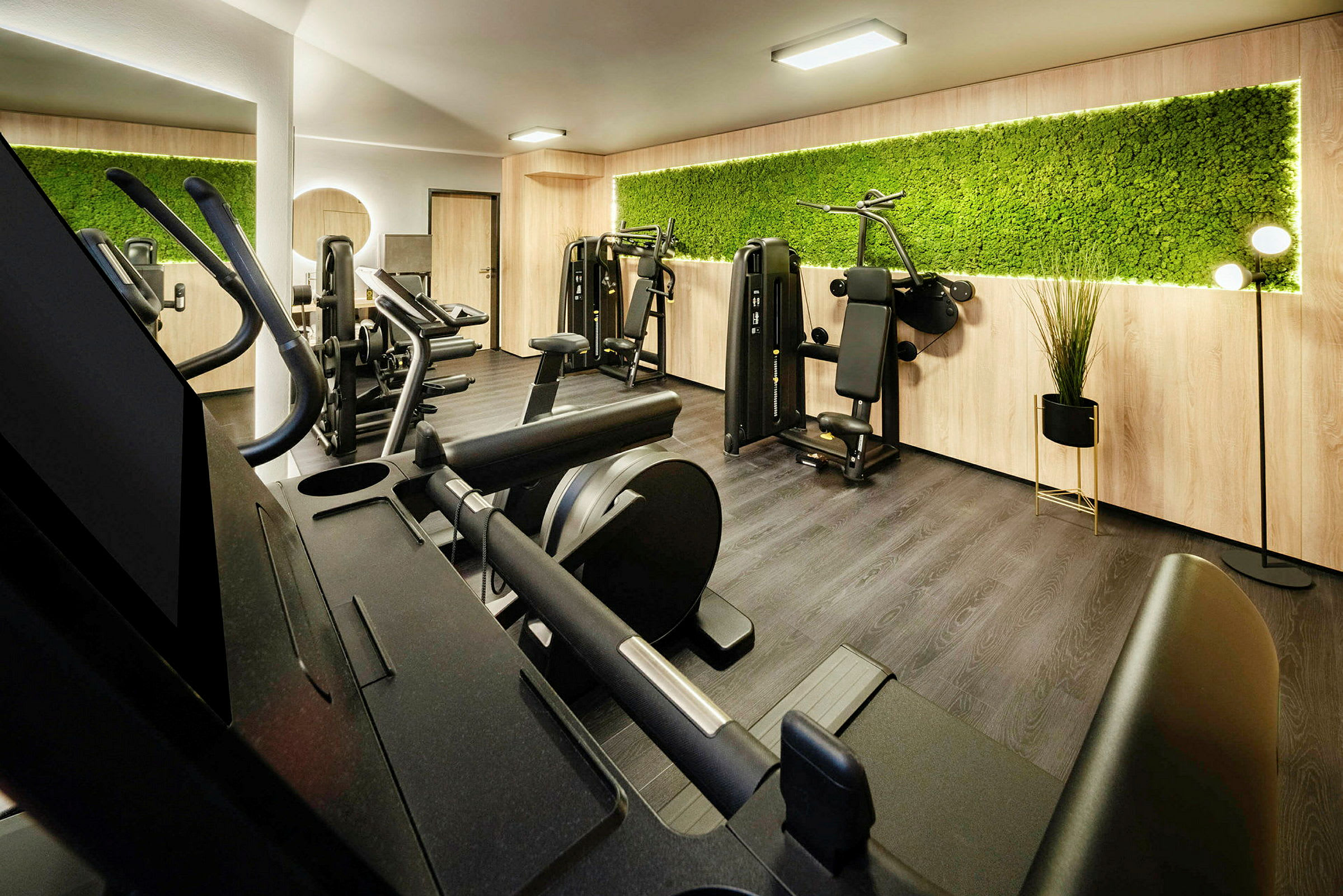 Ett gym med träningsmaskiner och cyklar. En grön vägg och trämaterial pryder interiören. (AI-genererad text)