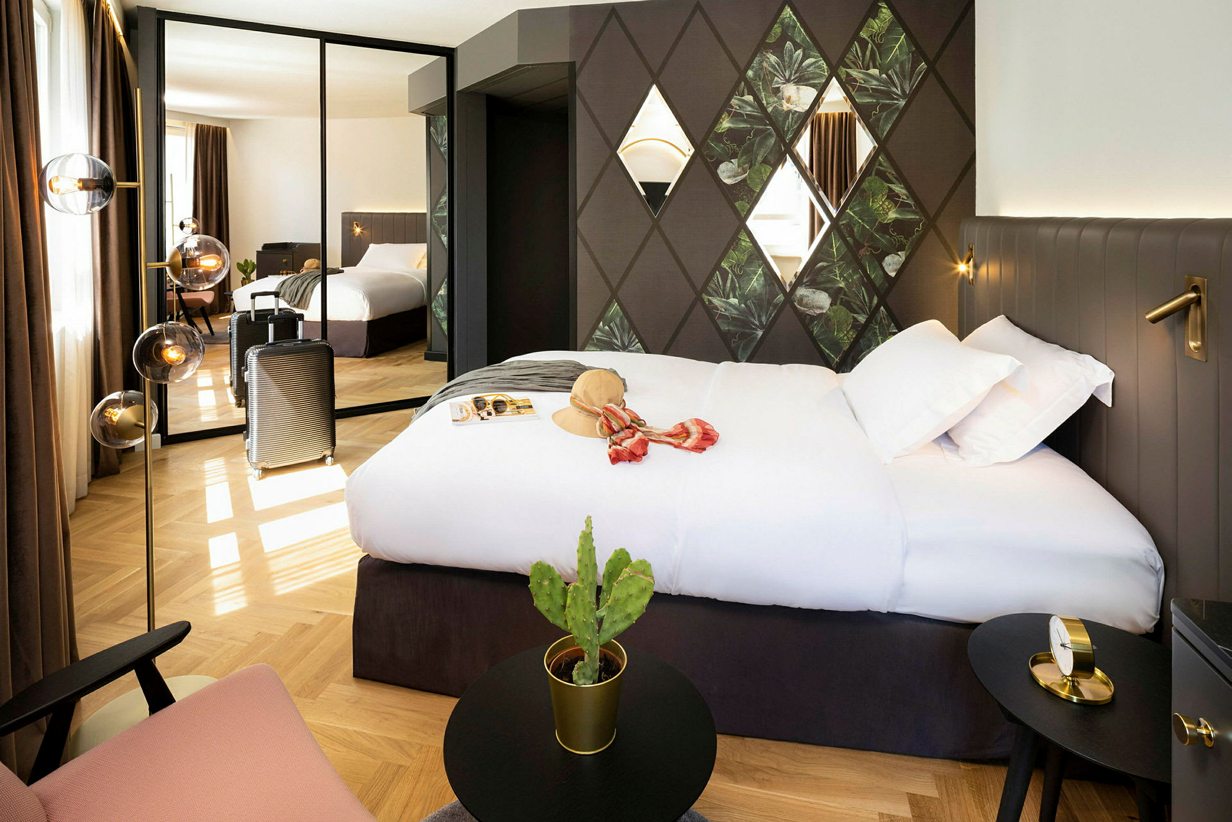 Ett hotellrum med en stor säng, väggdekorationer och en spegel som reflekterar rummet. En resväska står vid sidan och det finns en liten kaktus på ett bord. (AI-genererad text)