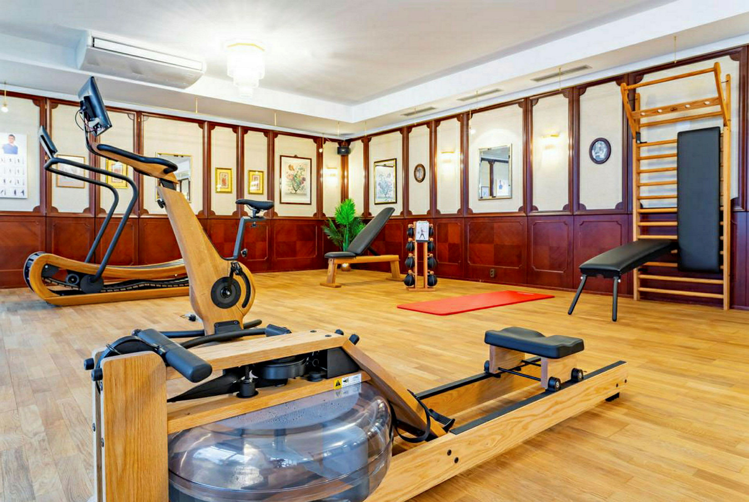 Bild på ett träningsrum med olika gymredskap som roddmaskin, motionscykel och träningsbänk. Rummet har träpaneler och konst på väggarna. (AI-genererad text)