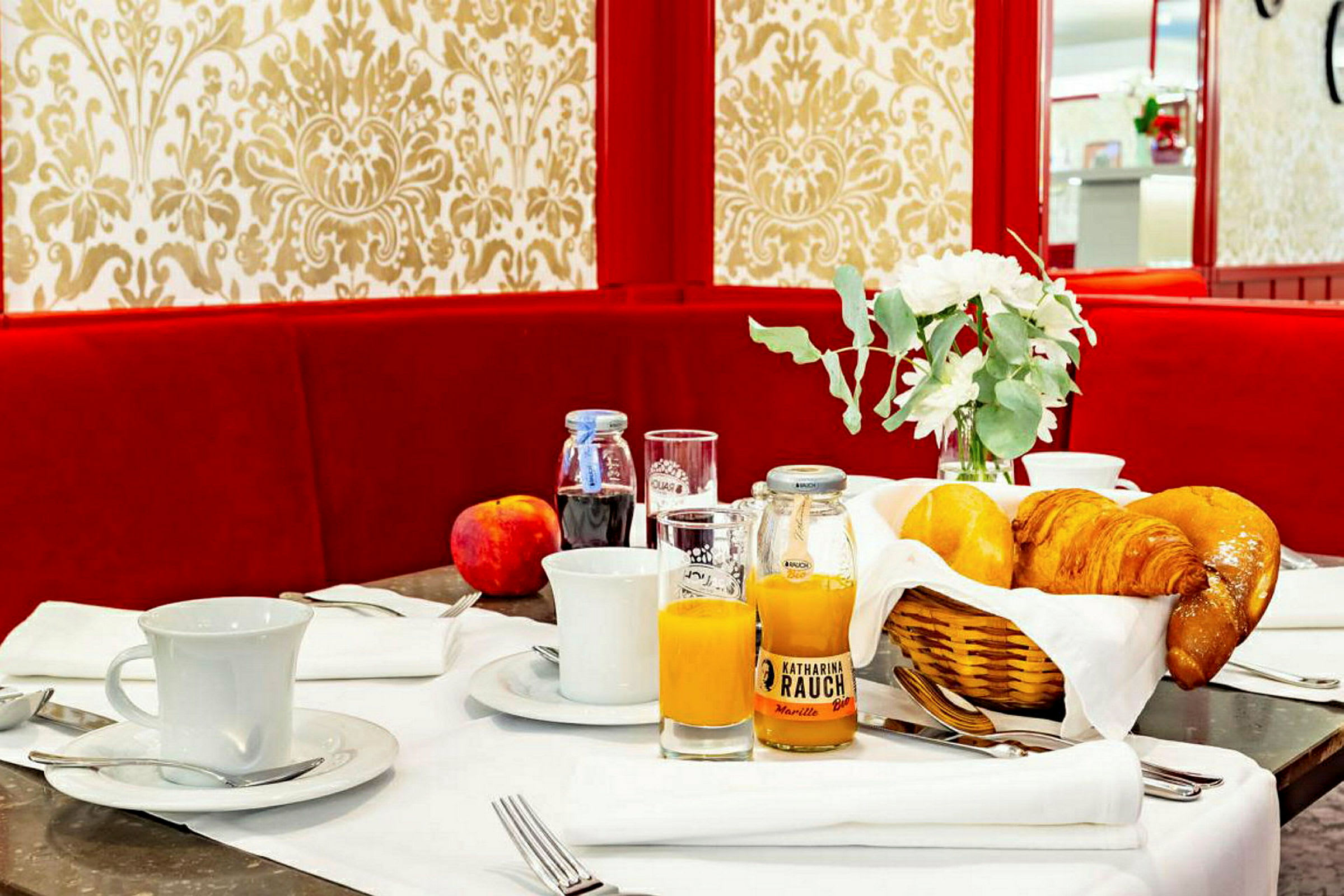 Frukostbord med bakverk, juice, kaffekoppar och dekorationer mot en rödfärgad soffa och mönstrad bakgrund. (AI-genererad text)