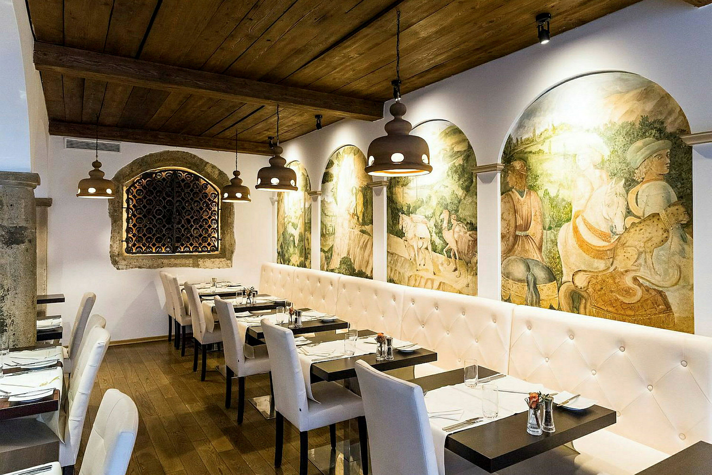 En restauranginteriör med vita stoppade soffor och stolar, mörka träbord samt en vägg dekorerad med historiska freskomålningar. Taket är av trä och belysningen består av hängande armaturer. (AI-genererad text)