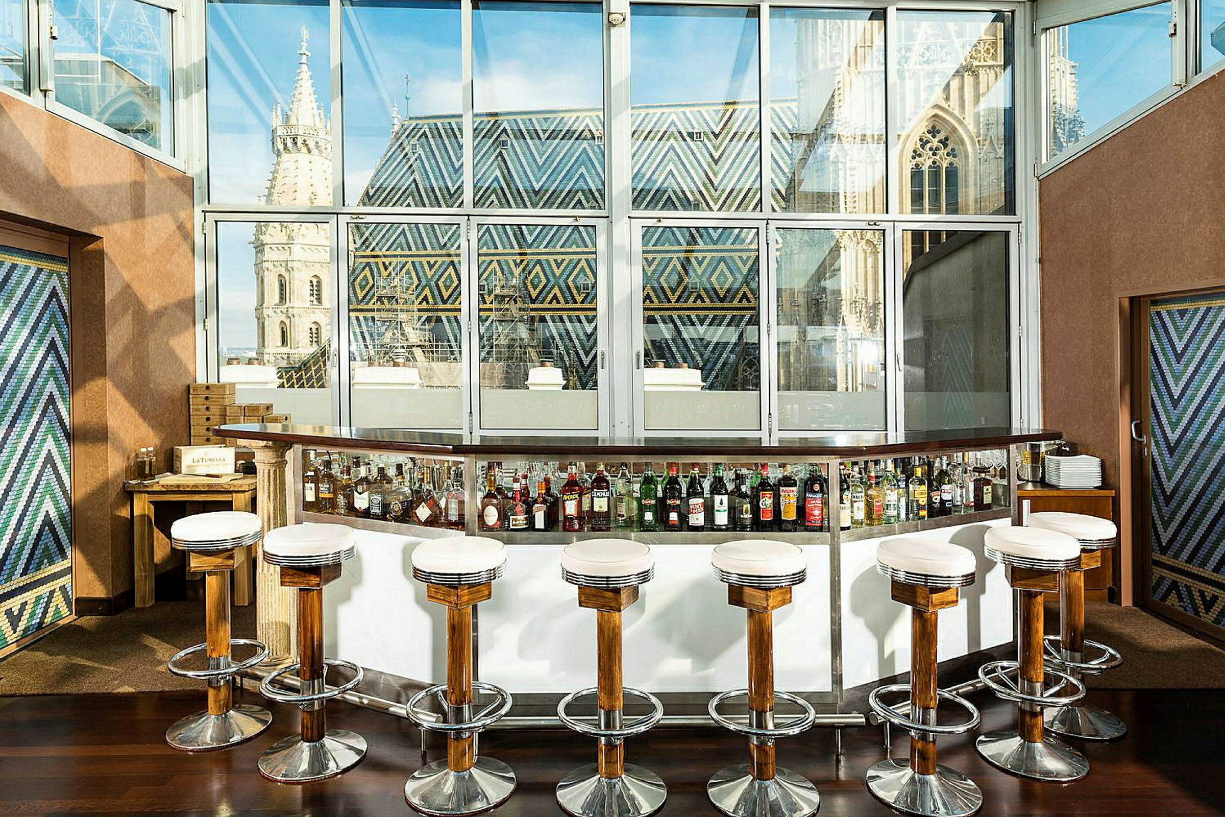 En cocktailbar med höga stolar framför en glasvägg med utsikt över en byggnad med färgglada takdekorationer. (AI-genererad text)