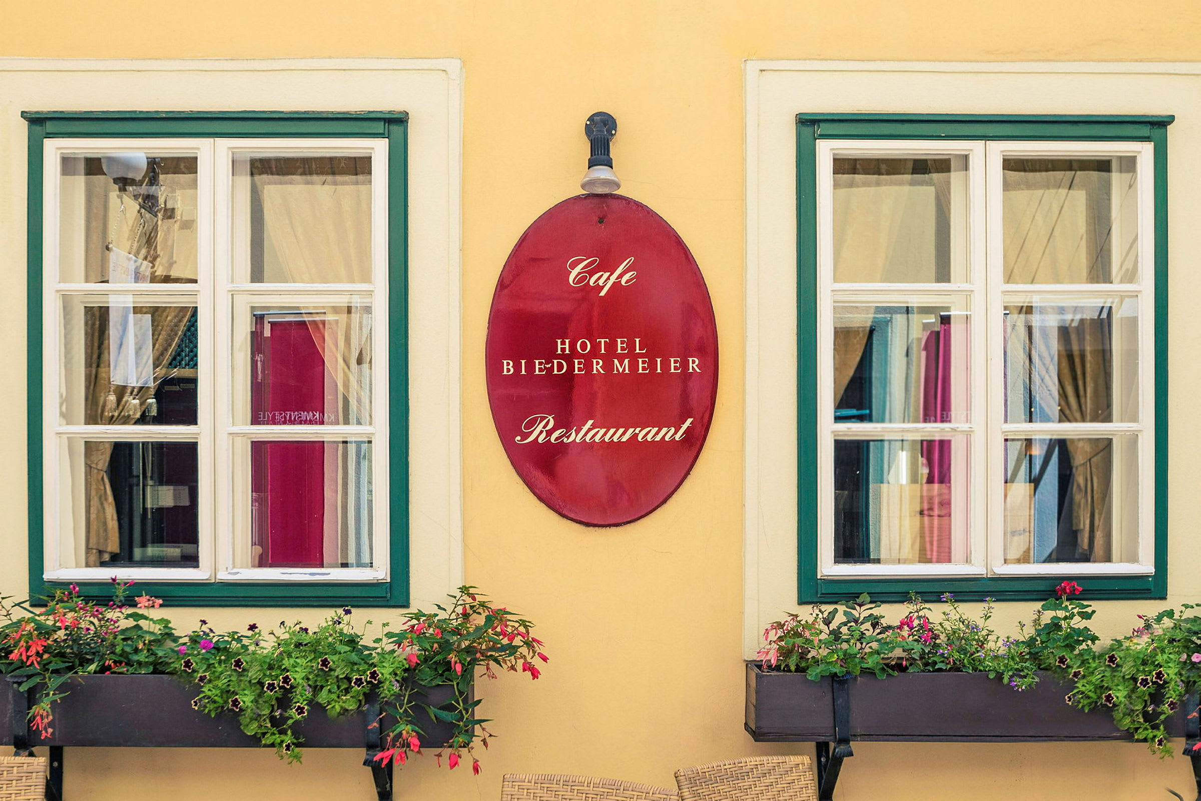 Två fönster med vita karmar och grön inramning, mellan dem hänger en röd skylt med texten "Cafe Hotel Biedermeier Restaurant". Framför fönstren finns blomboxar med blommande växter. (AI-genererad text)