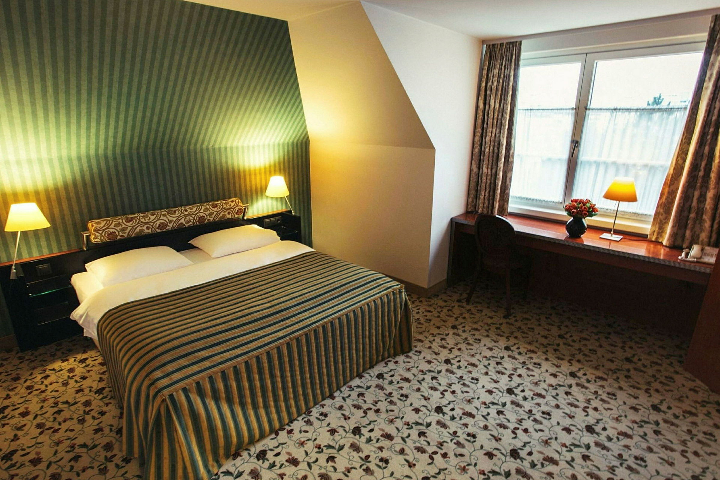 Ett hotellrum med en dubbelsäng, randigt överkast, två sängbord med lampor och en arbetsbänk med stol vid ett fönster. (AI-genererad text)