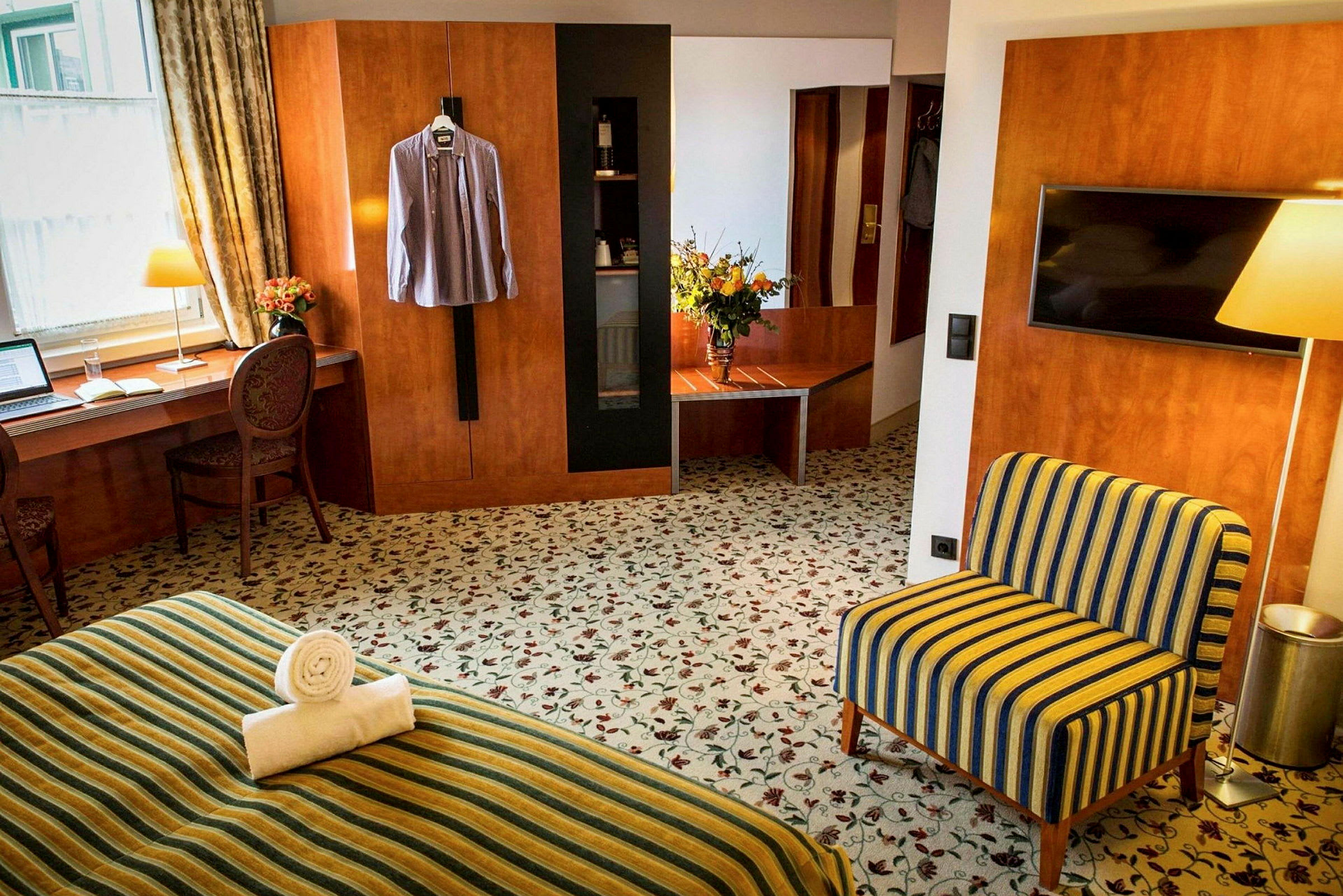 Ett hotellrum med ett skrivbord vid ett fönster, en stol, ett TV-skåp, en fåtölj och en säng klädd i randigt tyg. En skjorta hänger på väggen. (AI-genererad text)