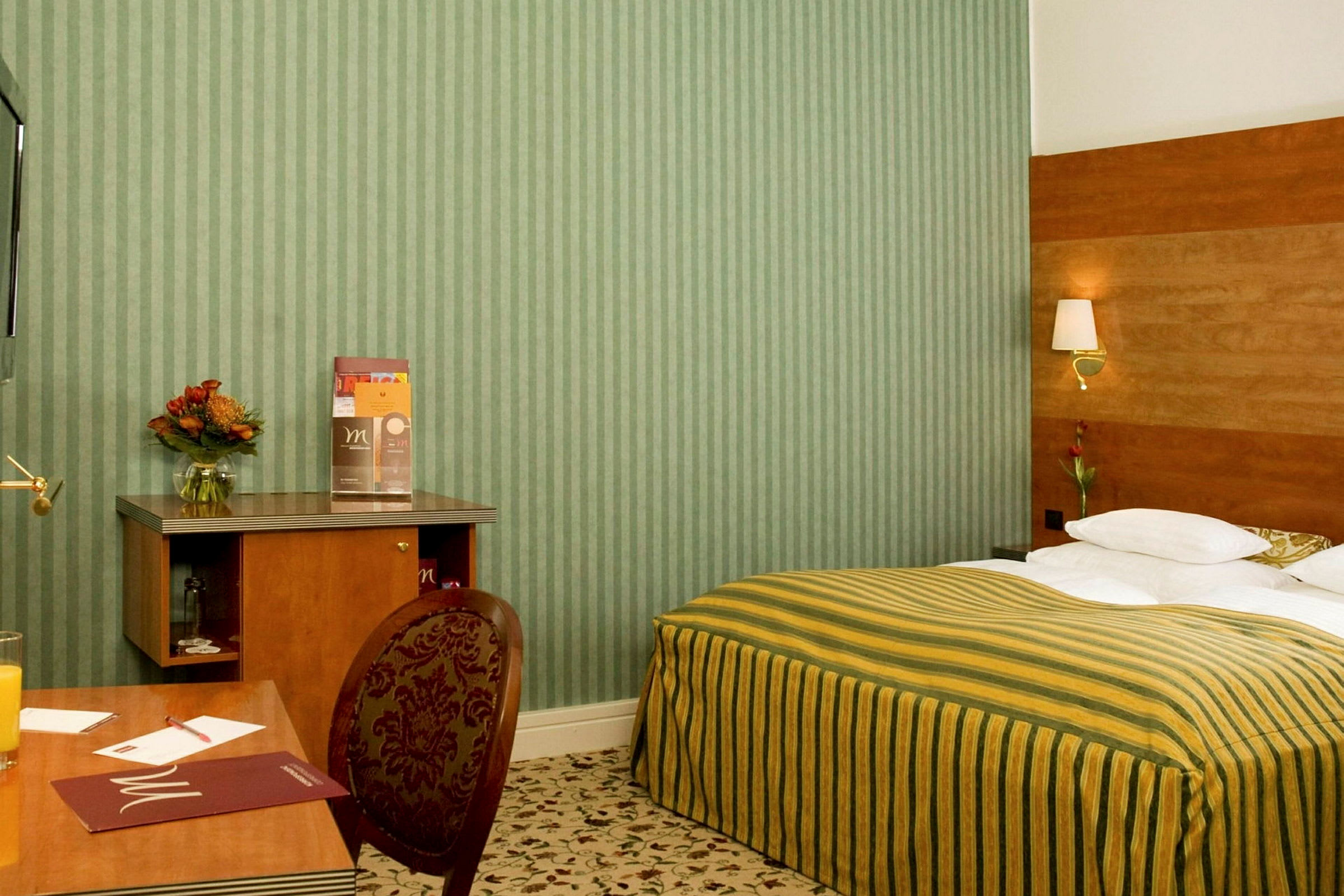 Ett hotellrum med en säng med randigt gulgrönt överkast, ett skrivbord och en stol. Det finns en blomvas och informationsmaterial på skrivbordet. (AI-genererad text)