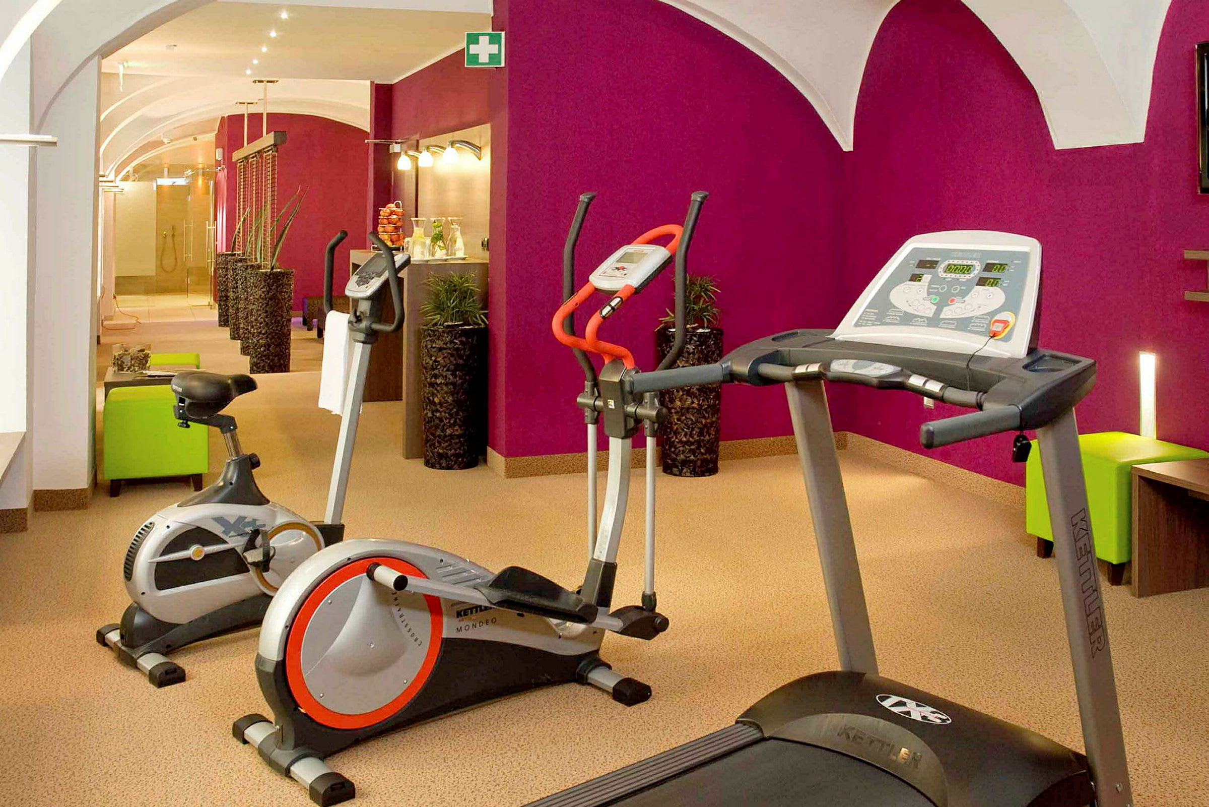 Ett gym med träningsmaskiner, inklusive löpband och motionscyklar, beläget i ett rum med en lila vägg och ljus inredning. (AI-genererad text)