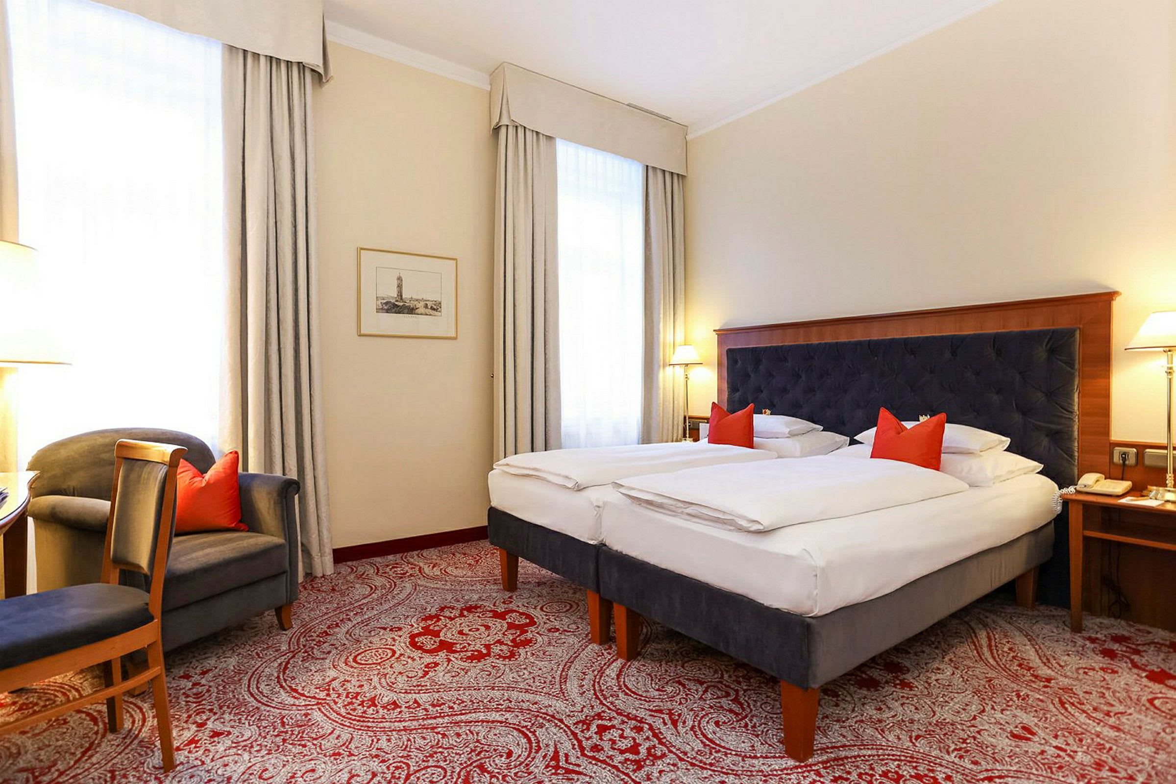 Ett hotellrum med två enkelsängar, ett mörkt klädd sänggavel, och röda prydnadskuddar. Rummet har vita väggar, stora fönster med ljusa gardiner och en röd mönstrad matta. (AI-genererad text)