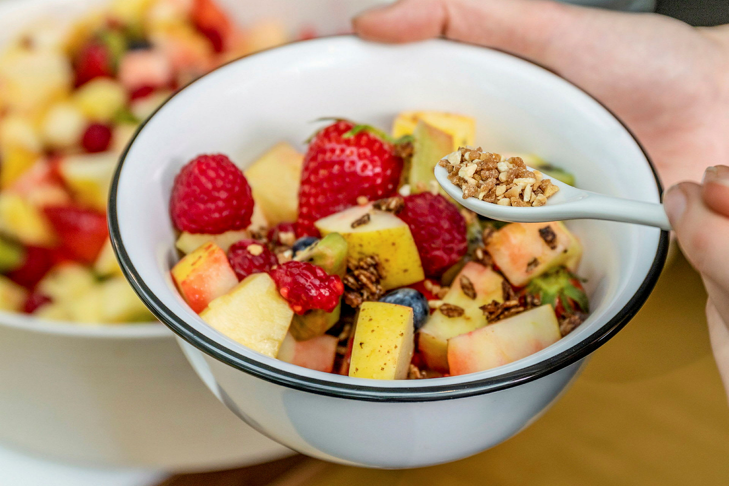 En skål med fruktsallad innehållande olika frukter och bär, samt en sked med granola. (AI-genererad text)
