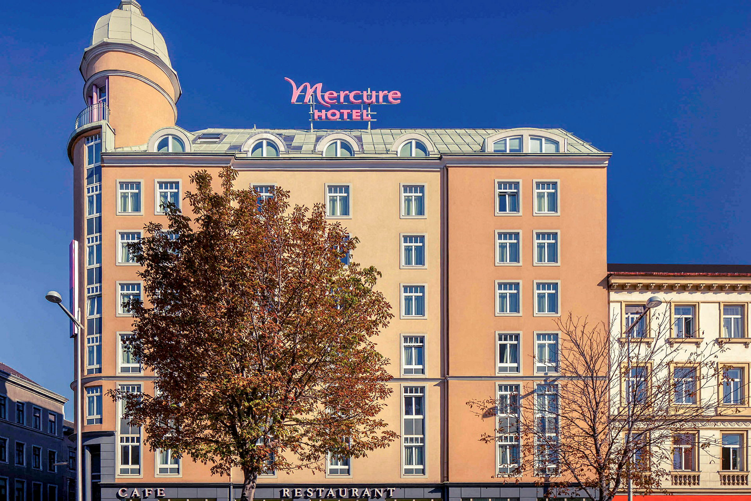 Exteriören av ett hotell med namnet Mercure, med en beige fasad, många fönster och ett högt torn upptill. (AI-genererad text)