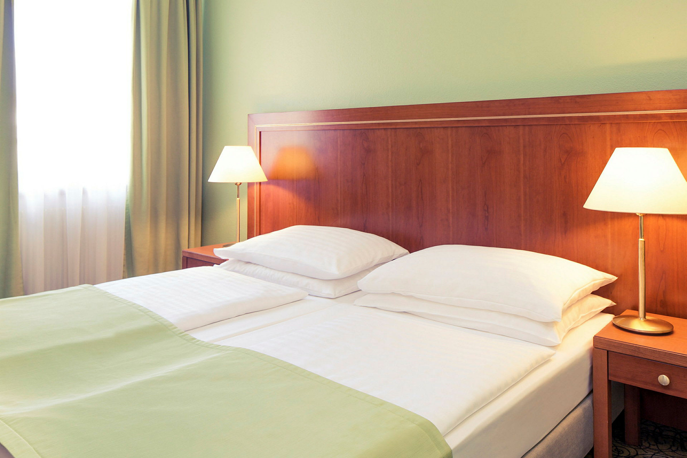 Ett hotellrum med en dubbelsäng som har vita sängkläder och gröna filtar. Två sänglampor står på träbord bredvid sängen. (AI-genererad text)