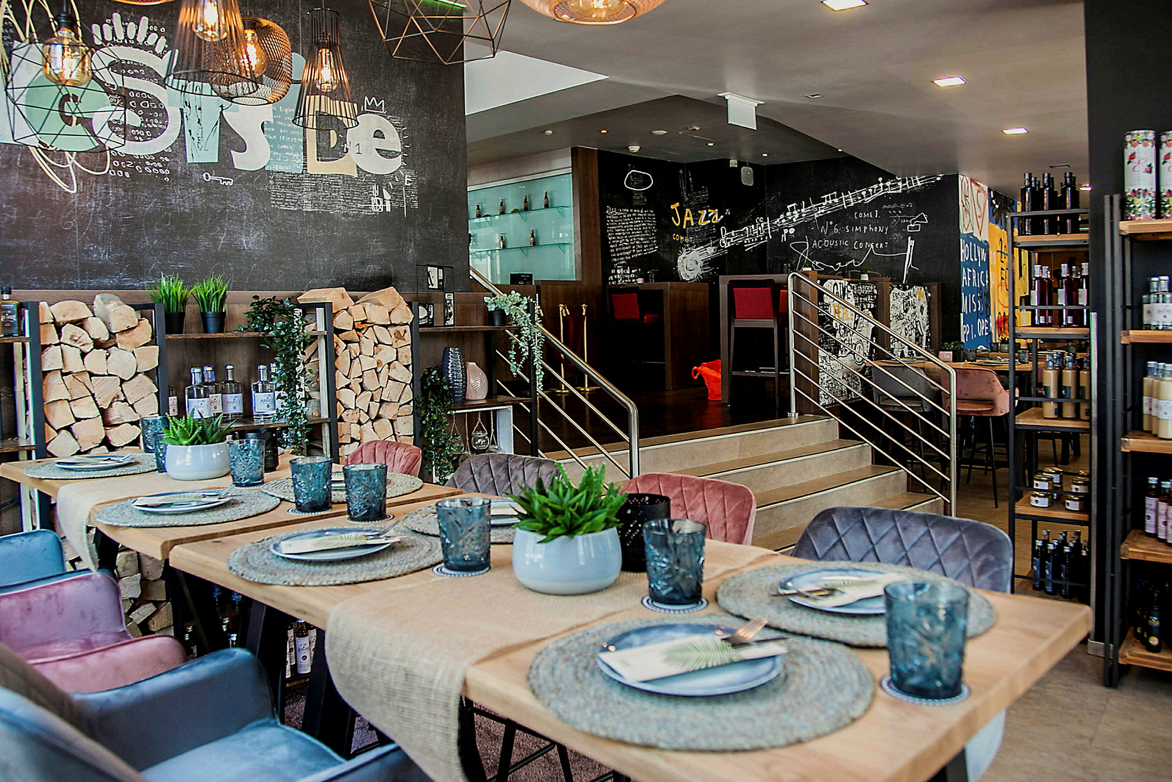 En restaurang eller cafe med ett modernt inredningsdesign. Bordet är dekorerat med tallrikar, glas och en liten växt. Väggarna har kreativa målningar och det finns trästaplar som en del av inredningen. (AI-genererad text)