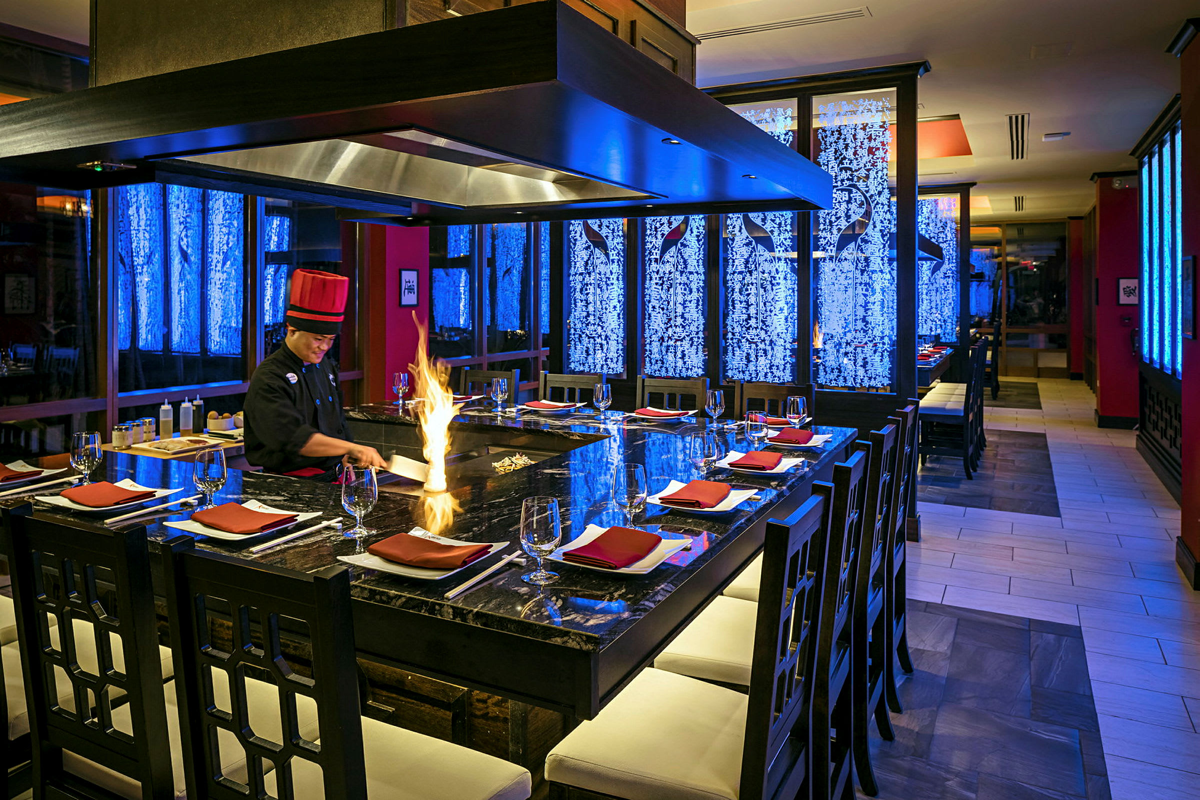 Restaurang med teppanyaki-kock vid en stor matlagningsstation, omgiven av stolar och dukning. Eldflammor syns på grillen. (AI-genererad text)