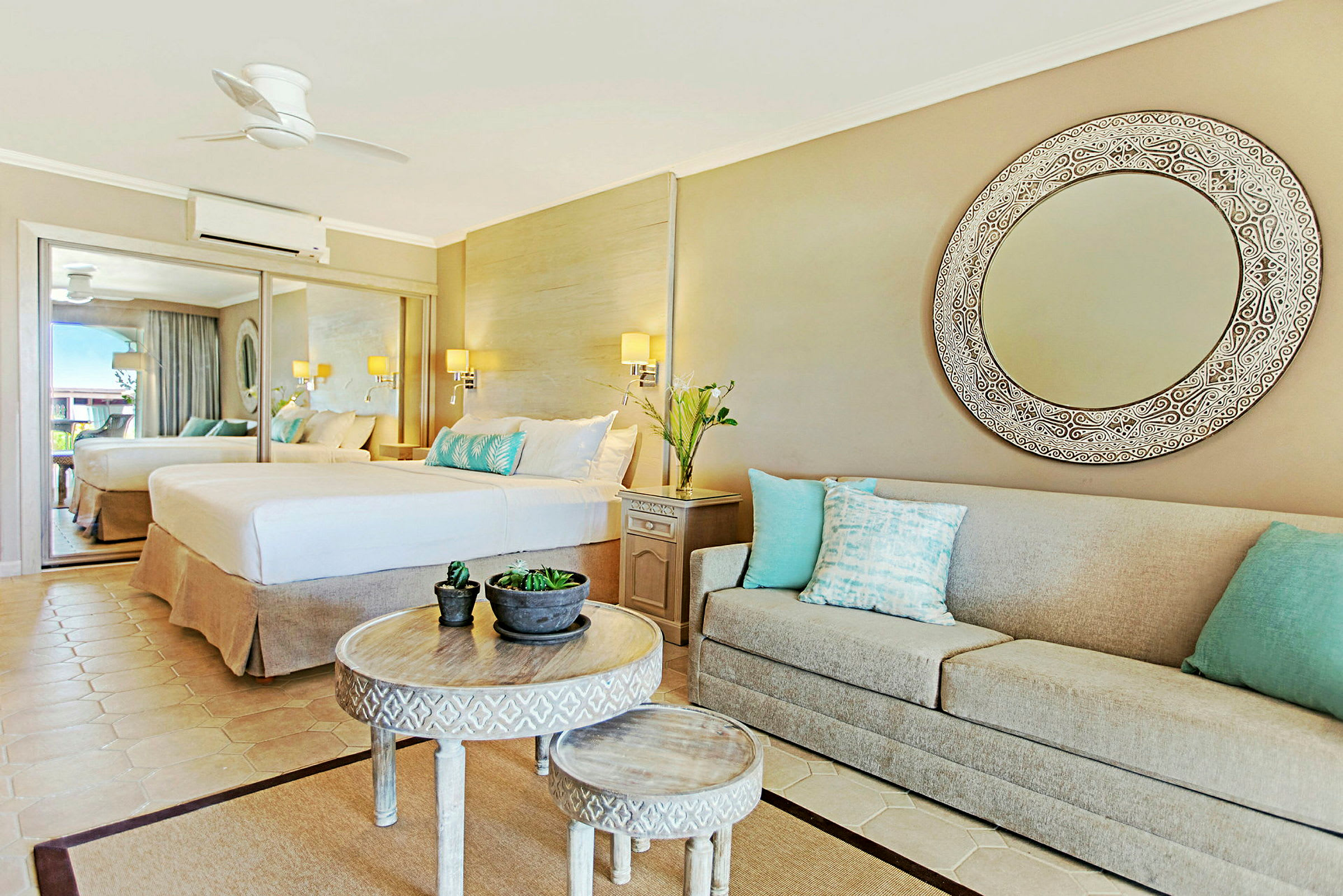 Ett elegant hotellrum med en stor spegel, en bred säng och en beige soffa med dekorativa kuddar. Ett runt bord med mindre bord och växtdekorationer i mitten. (AI-genererad text)
