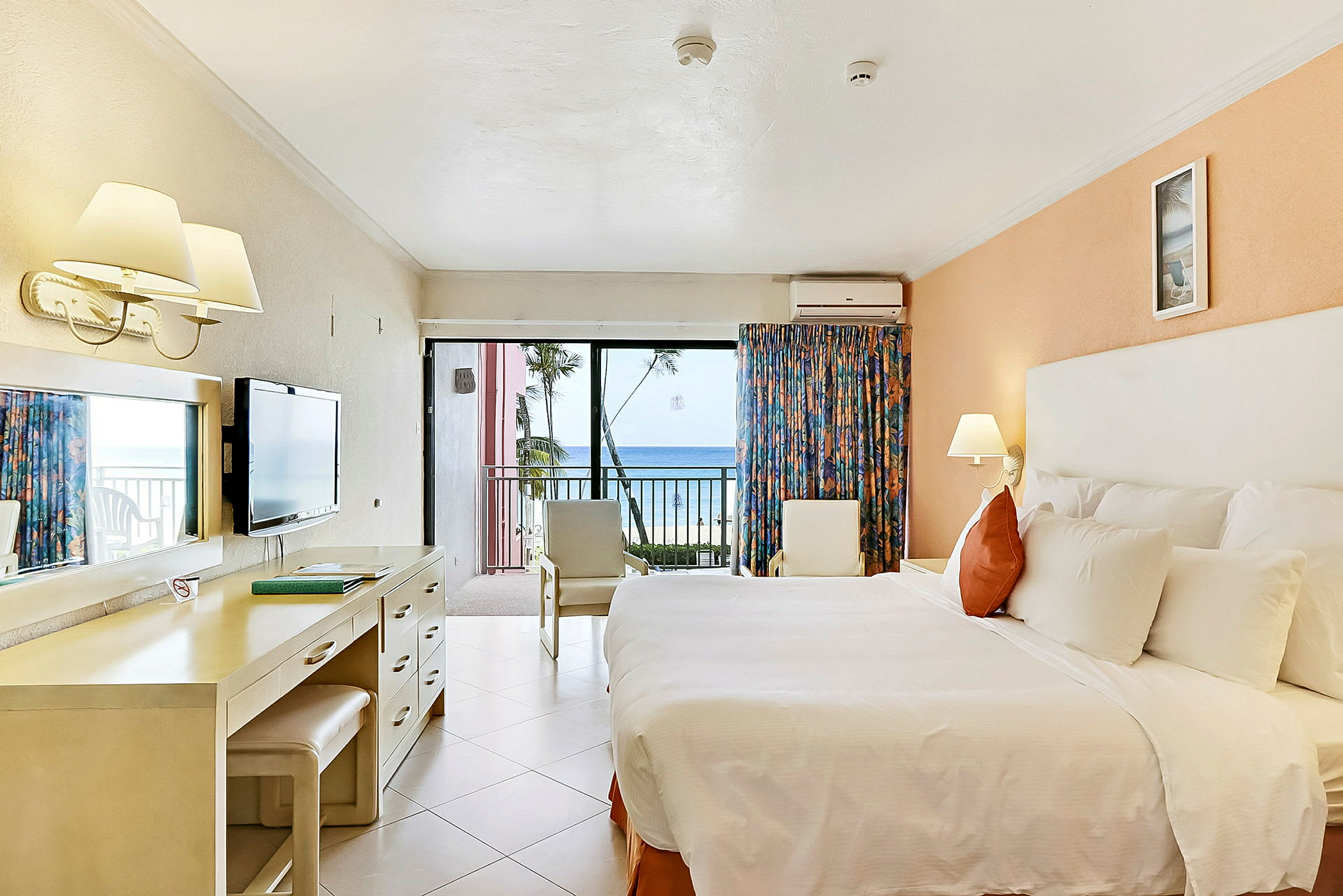 Hotellrum med en stor säng, skrivbord, stolar och en balkong med utsikt över havet. (AI-genererad text)