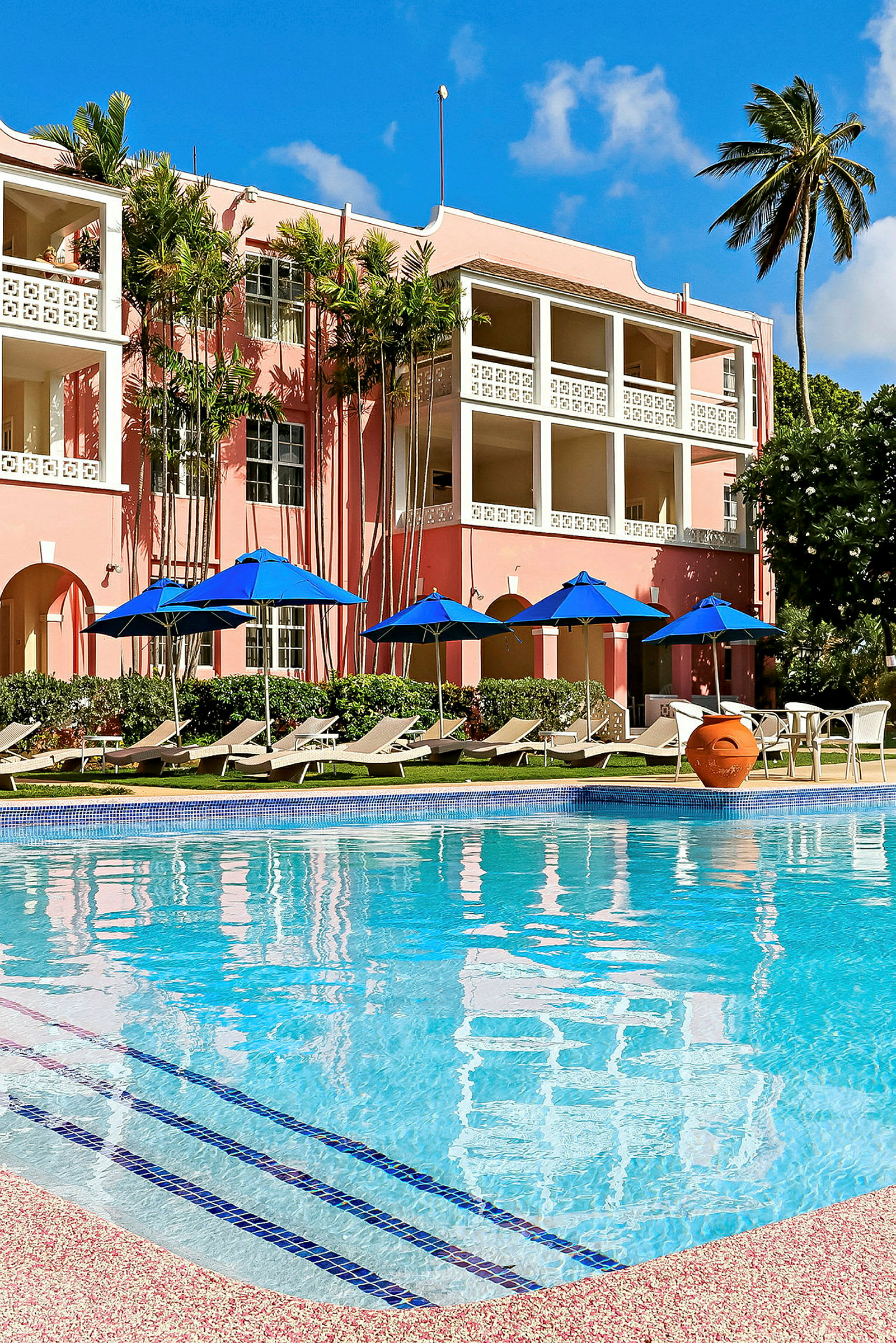 En pool framför ett rosa hotell med blå parasoller och solstolar i en tropisk miljö. (AI-genererad text)
