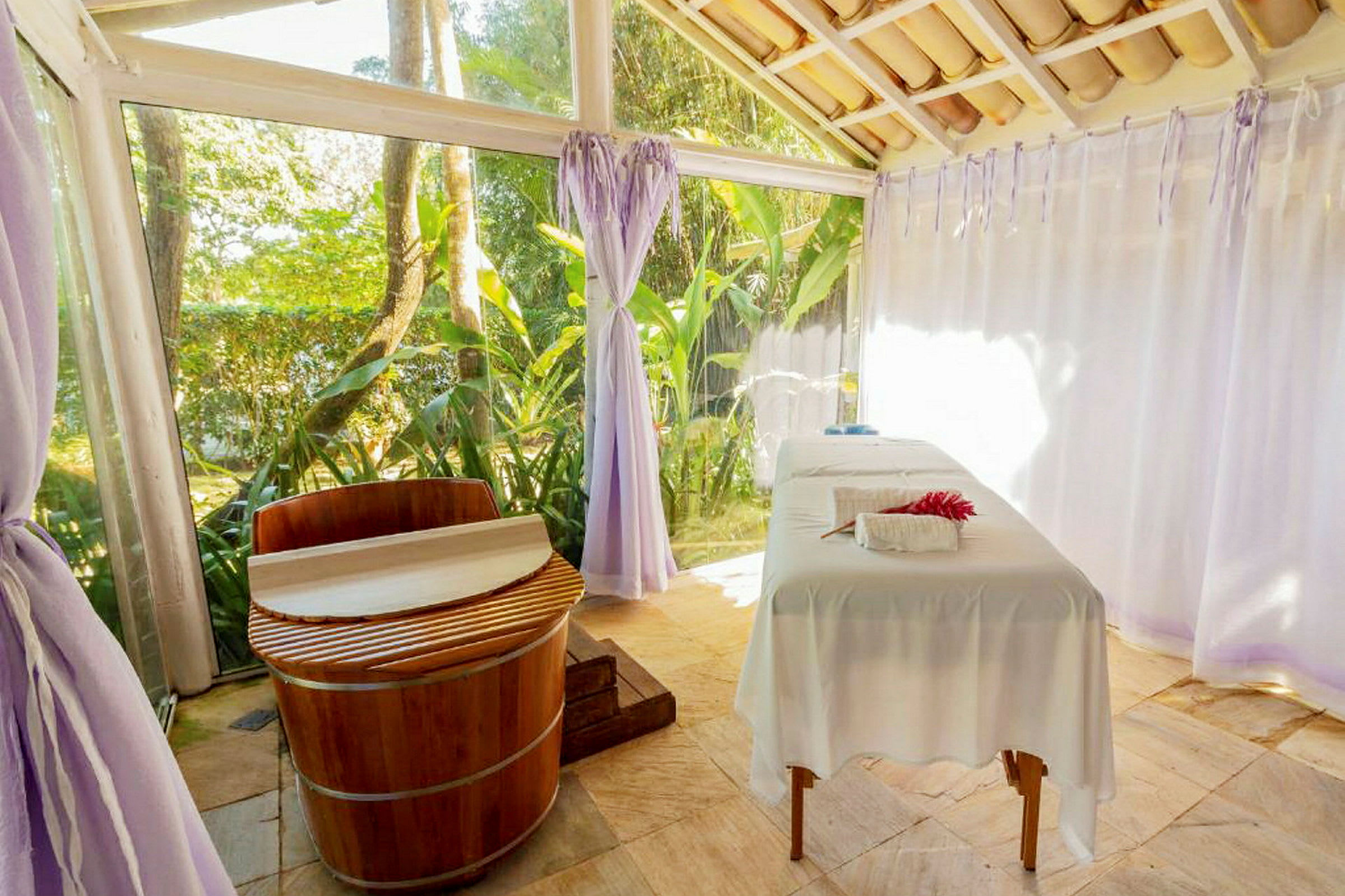 Ett spa-rum med en massagesäng och ett träbadkar, omgiven av stora fönster och gardiner samt grönska utanför. (AI-genererad text)