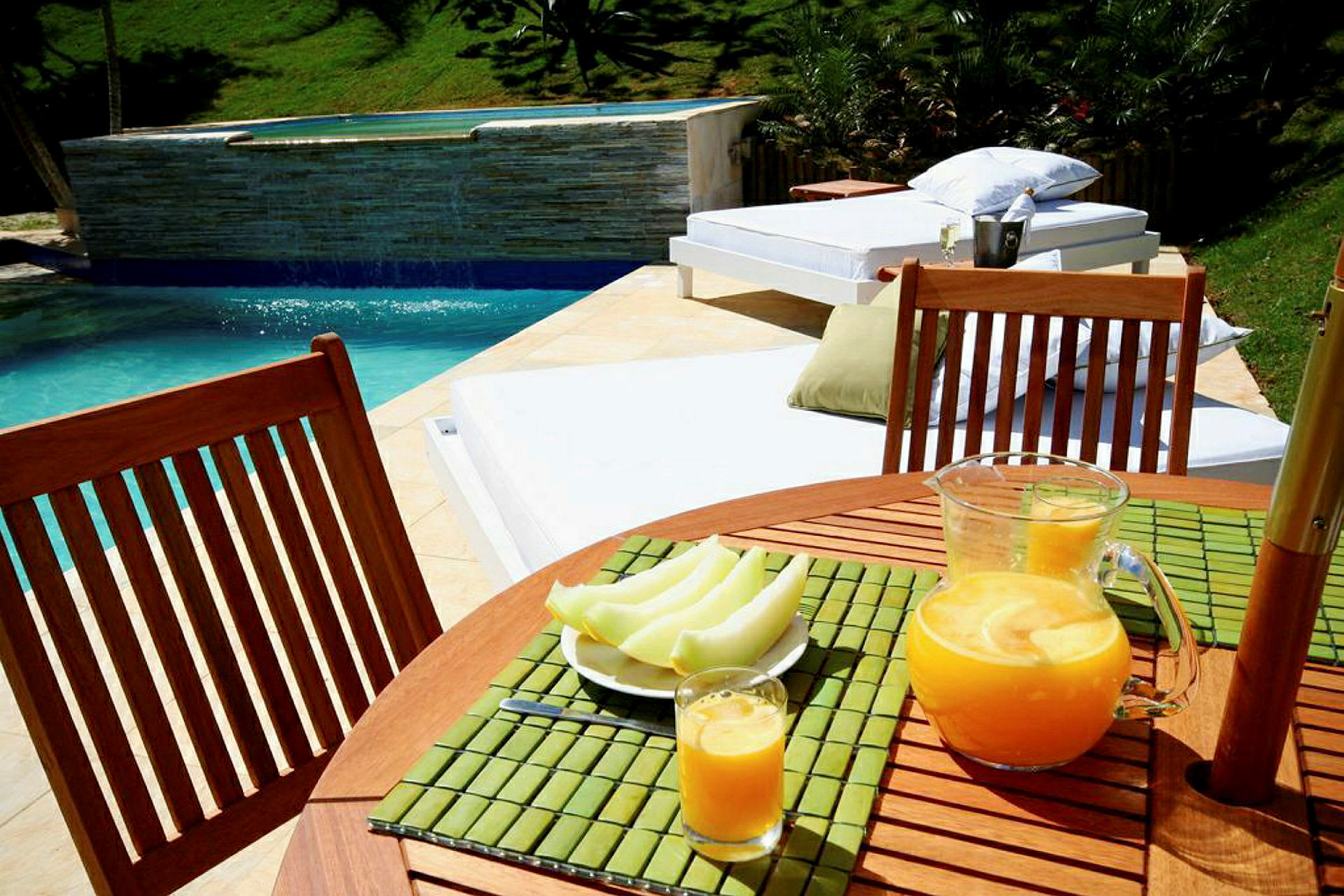 På bilden syns ett bord vid en pool, med en kanna och ett glas juice samt en tallrik med uppskuren frukt. I bakgrunden ligger solsängar och gröna växter. (AI-genererad text)