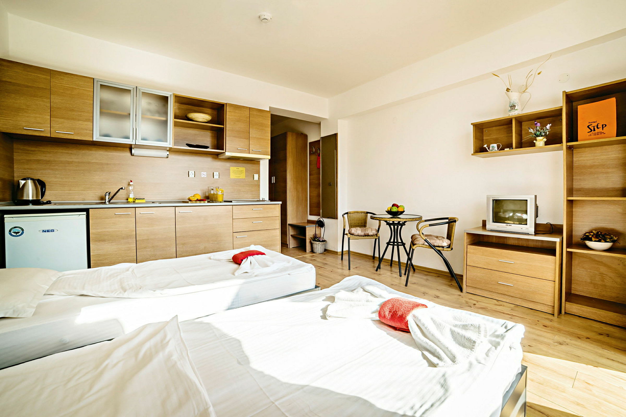 Ett hotellrum med två enkelsängar, en köksdel med skåp, samt ett litet bord och stolar. (AI-genererad text)