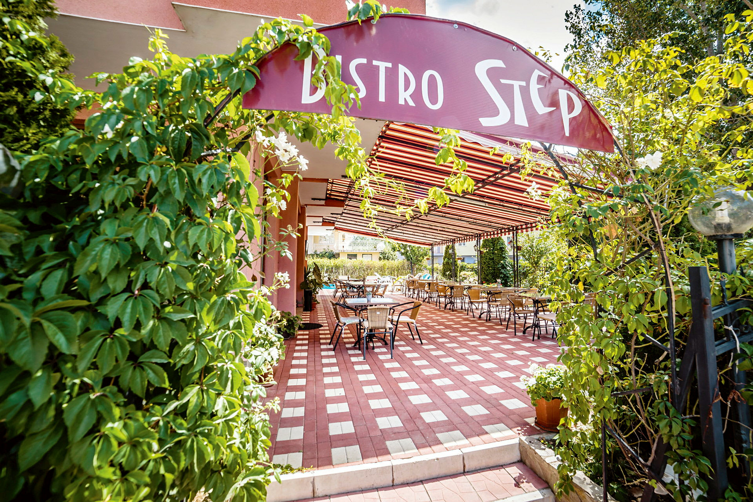 Ingång till en restaurang med en skylt "Bistro Step", omgiven av grönska och med uteservering under tak. (AI-genererad text)