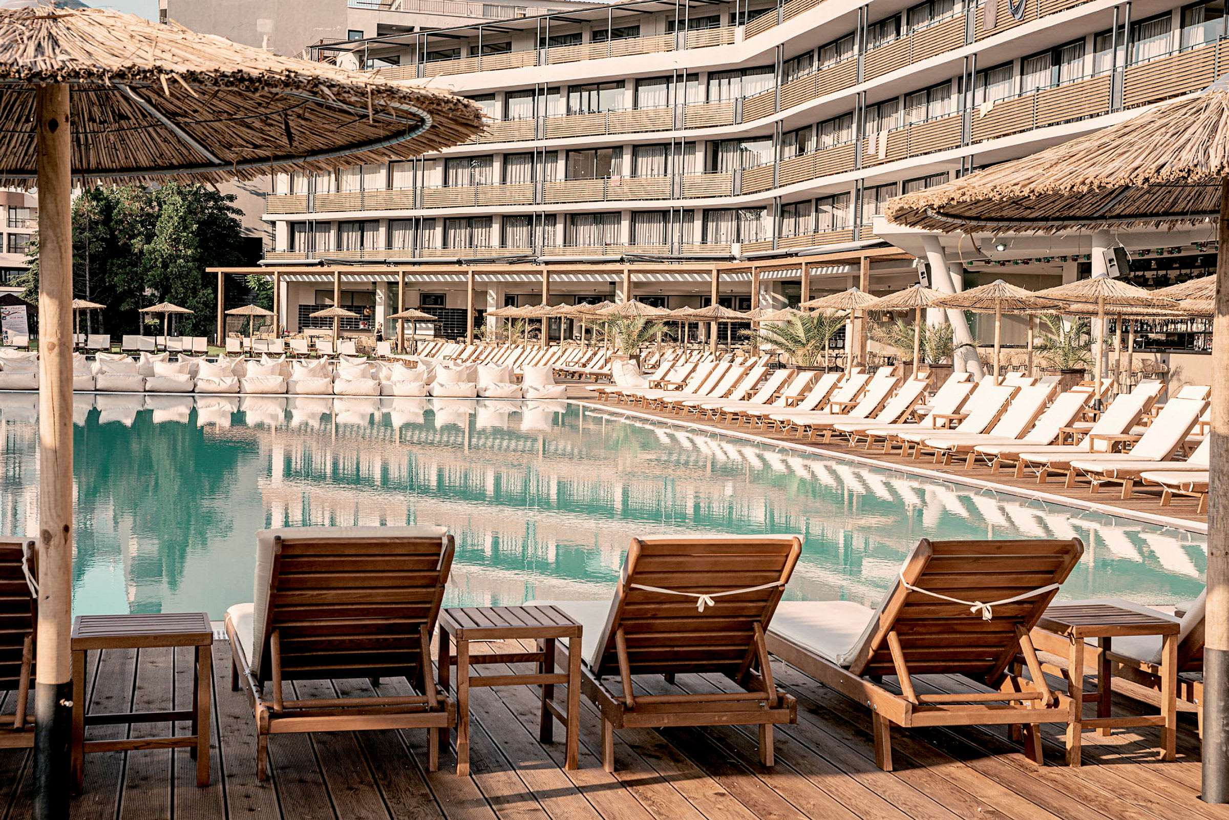 Stolar och bord står vid en pool, omgivna av parasoller och ett hotell i bakgrunden. (AI-genererad text)