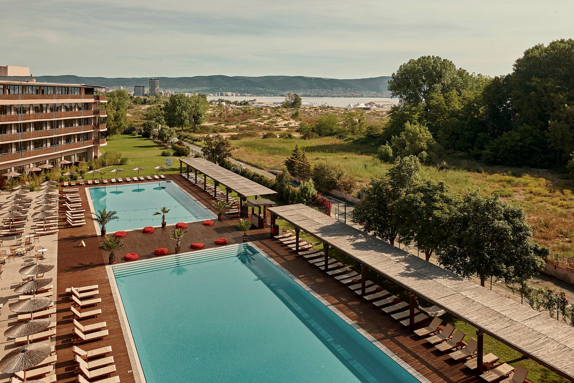 Stor pool med solstolar och parasoller vid sidan av ett hotell, med grönska och landskap i bakgrunden. (AI-genererad text)