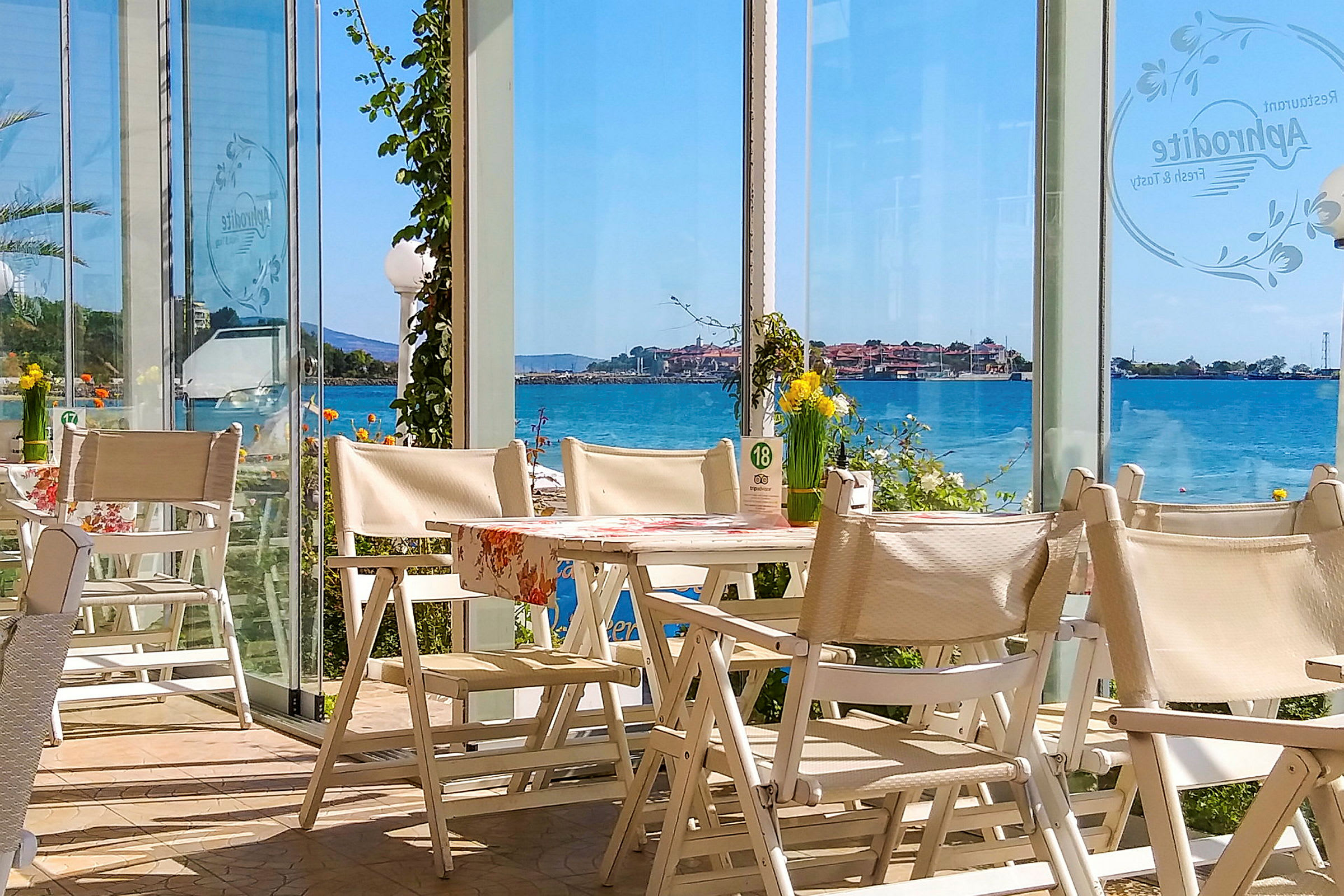 Utsikt från en restaurang med vita stolar och bord, med glasväggar som erbjuder vyer över havet och en kuststad. (AI-genererad text)