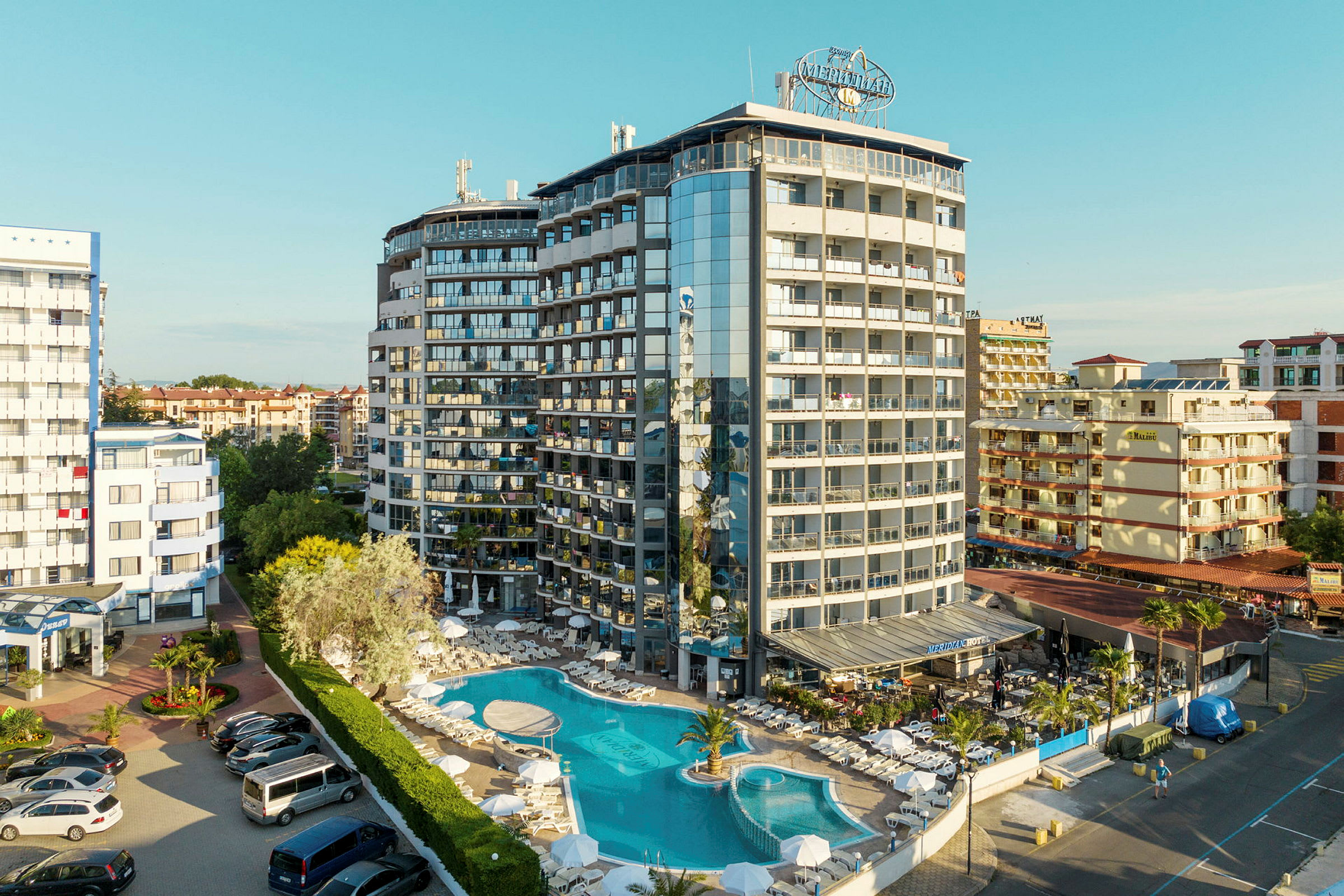Ett flervåningshotell med pool och solstolar framför, inbäddat i ett tätbebyggt område. (AI-genererad text)