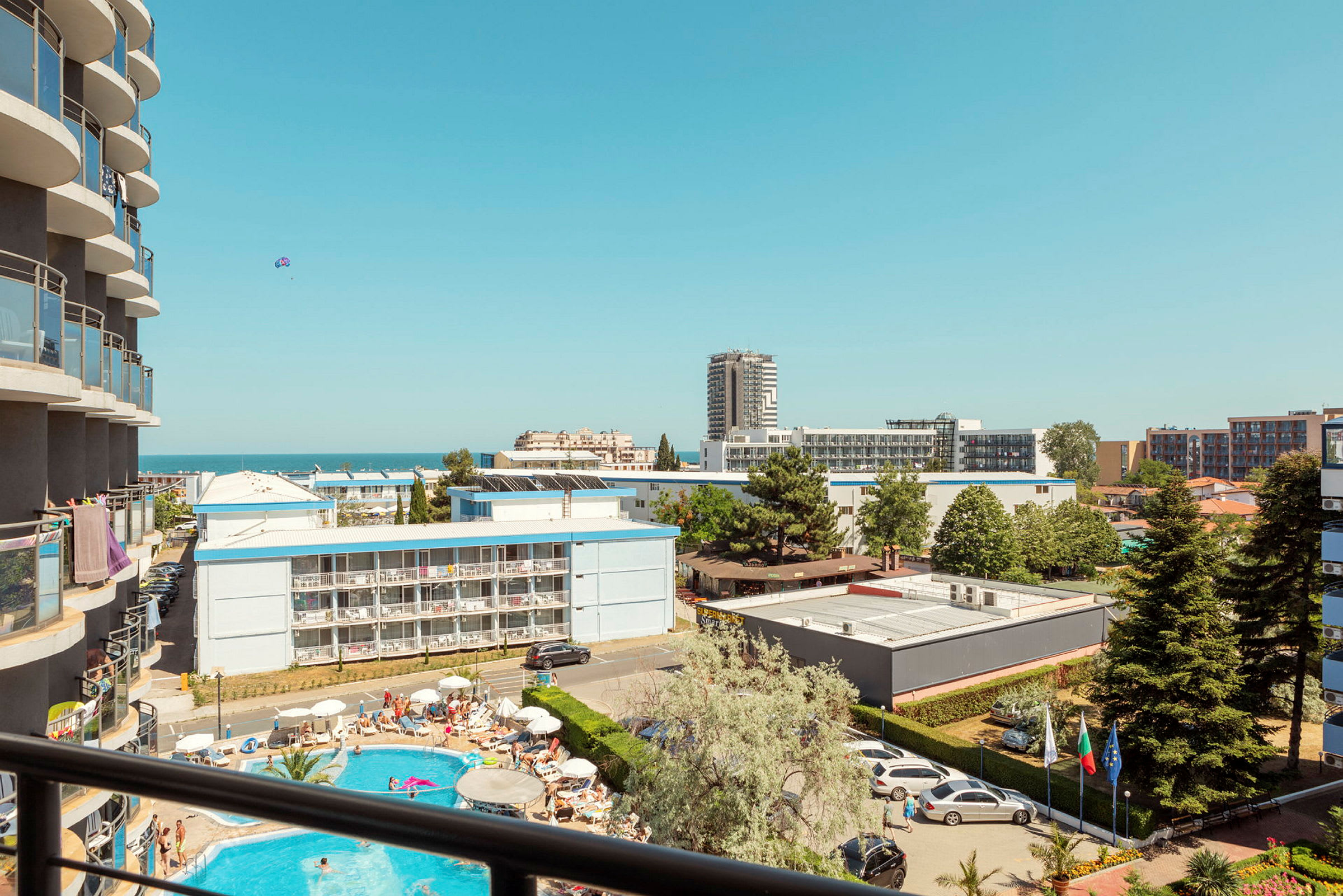 Balkong med utsikt över hotellområde, pool och byggnader, med havet i bakgrunden under klar himmel. (AI-genererad text)