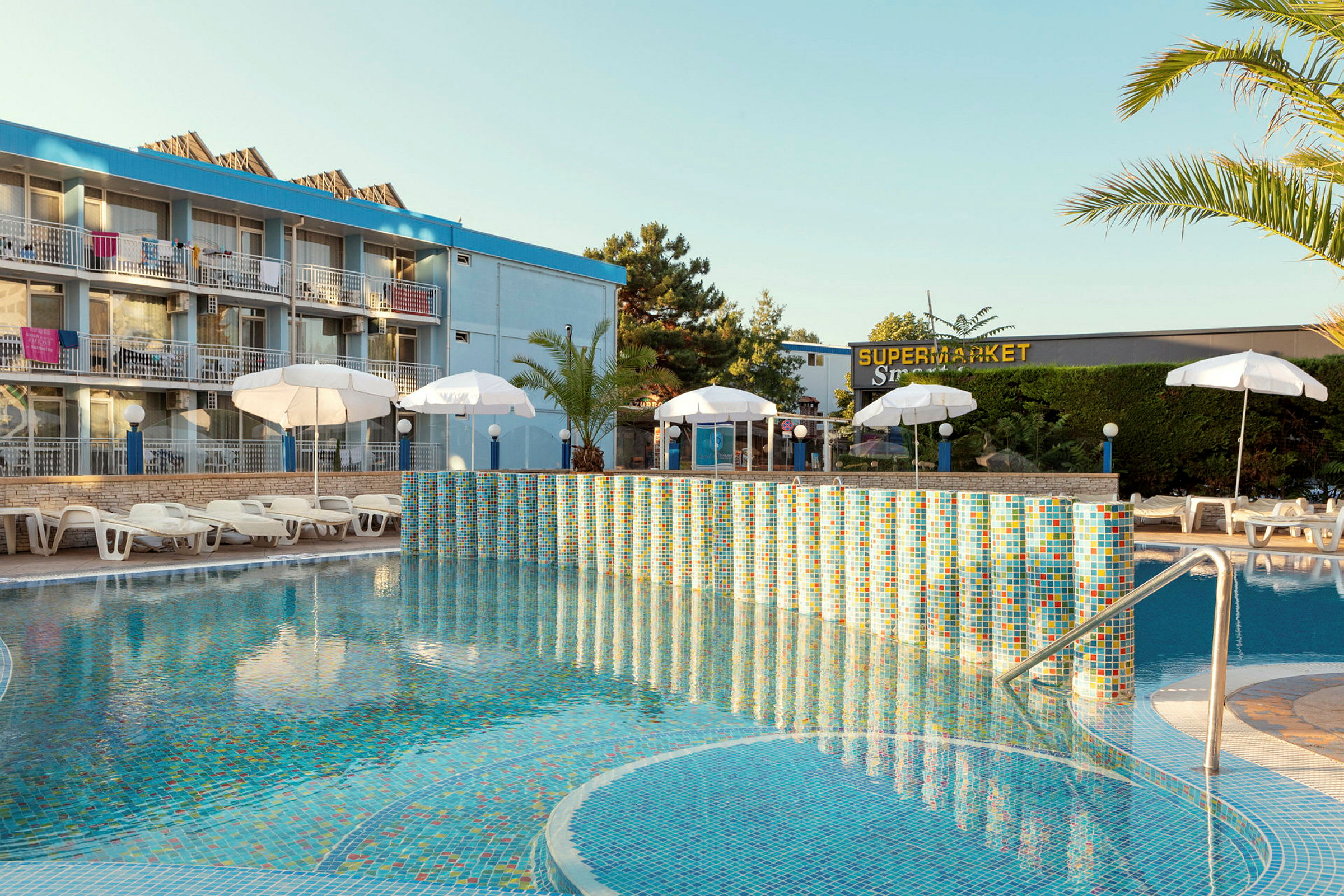 Hotell med pool, solstolar och parasoller framför byggnaden. (AI-genererad text)