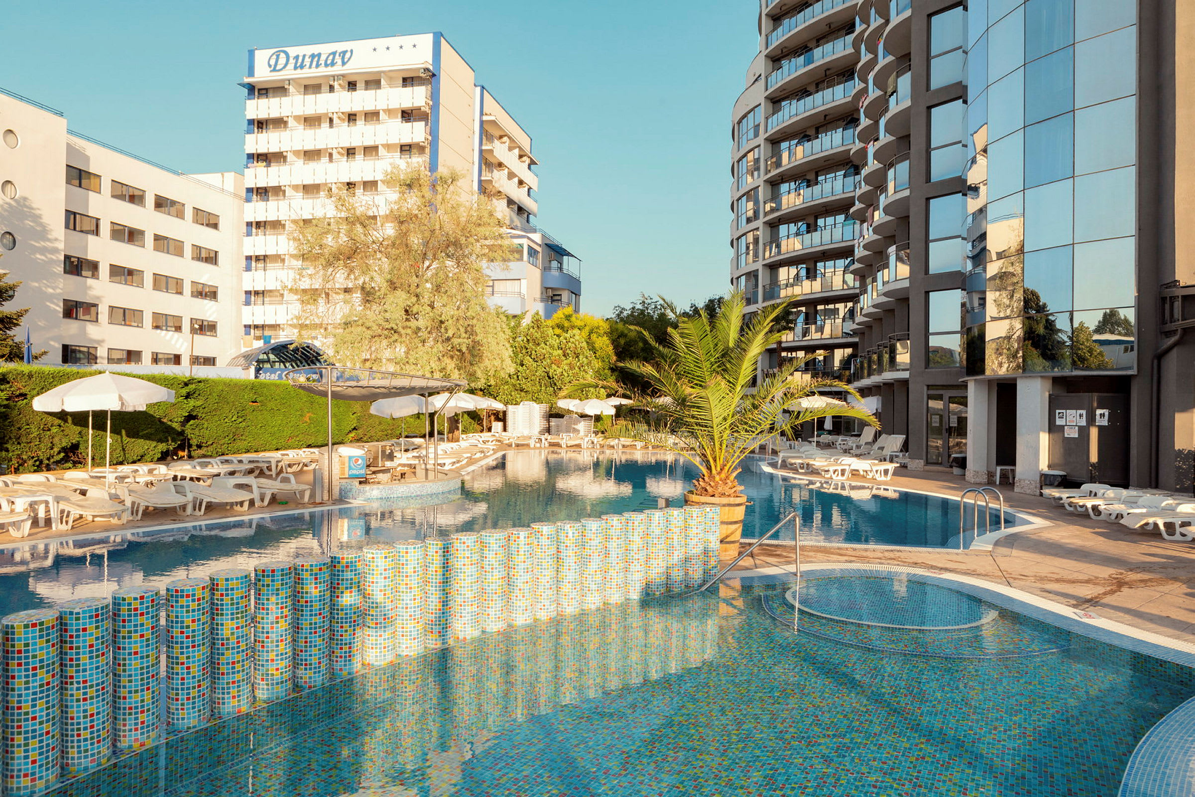 Bild av hotellområde med utomhuspool, omgivna av solstolar och parasoller. Bakgrund med byggnader och växtlighet. (AI-genererad text)