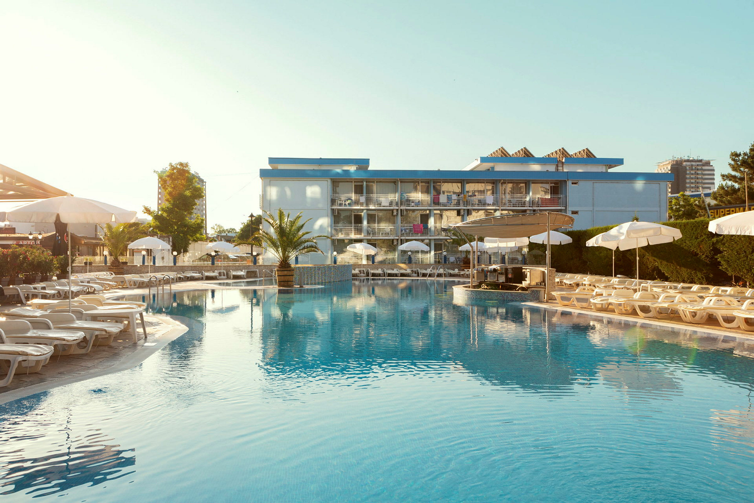 En pool framför ett hotell med solstolar och parasoller runt omkring. (AI-genererad text)