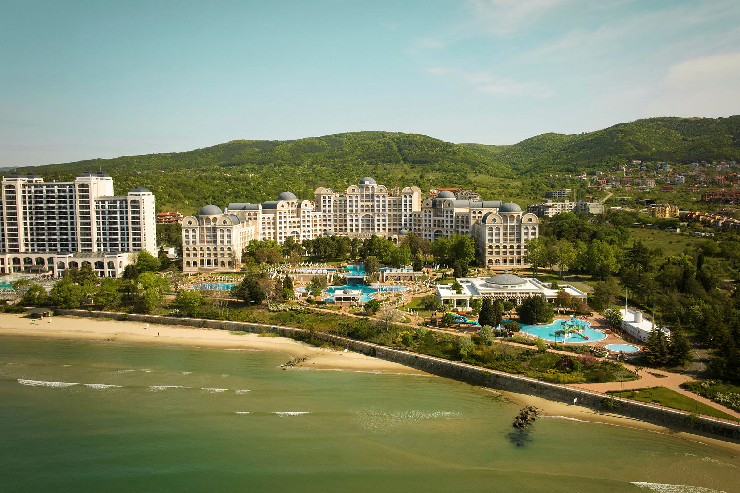 Vy över hotellkomplex vid stranden med pooler och grönskande omgivning. (AI-genererad text)
