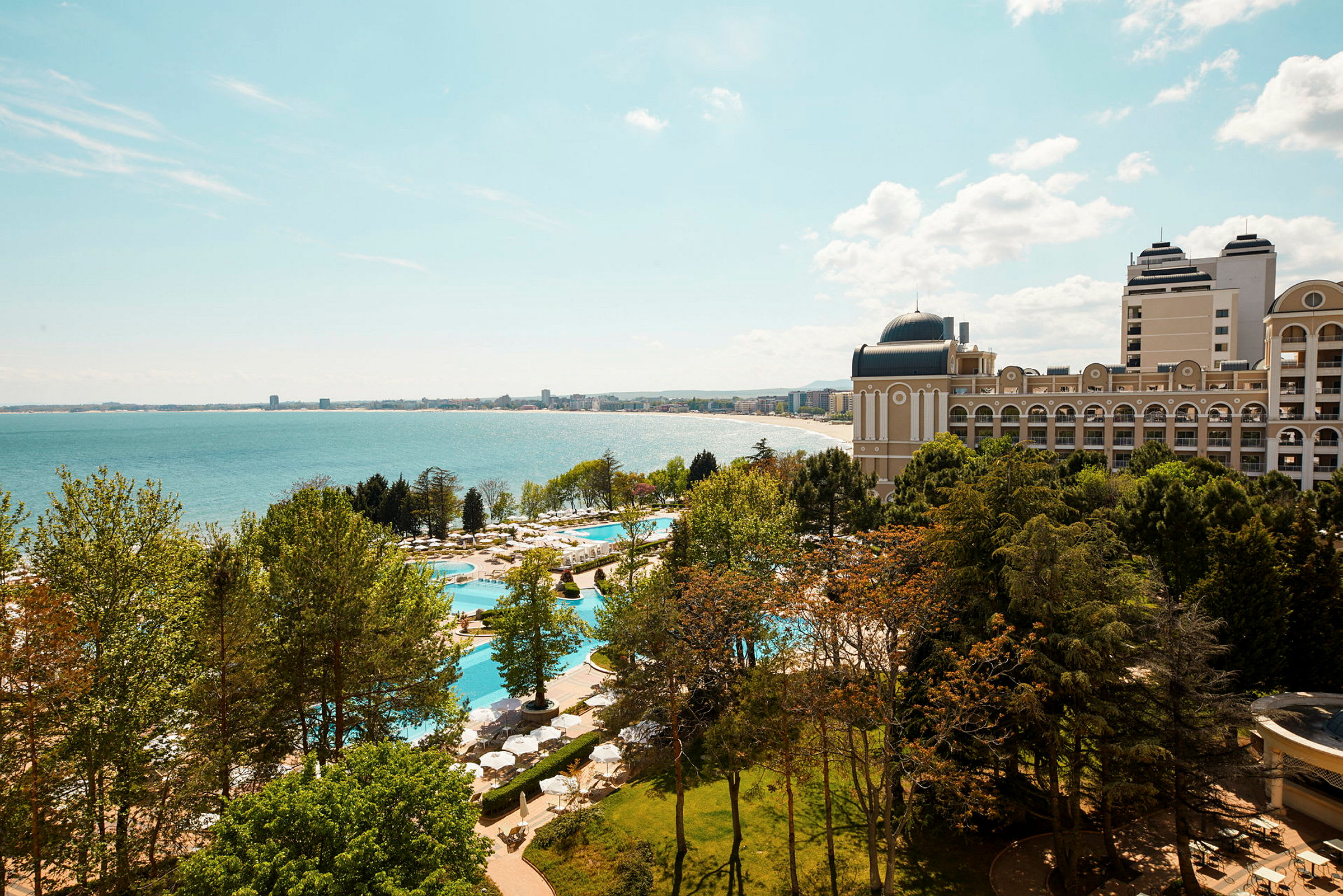 Bild av ett hotell med poolområde omgivet av grönskande träd, med utsikt över havet och en strand i bakgrunden. (AI-genererad text)