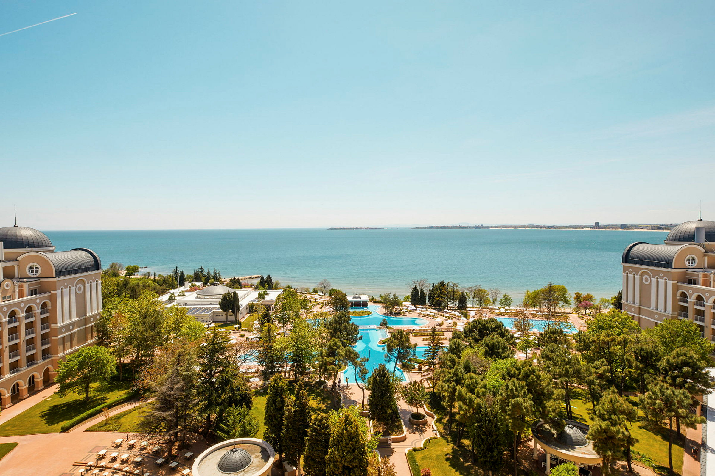 Utsikt över ett hotellområde med två byggnader, pooler, grönska och havet i bakgrunden. (AI-genererad text)